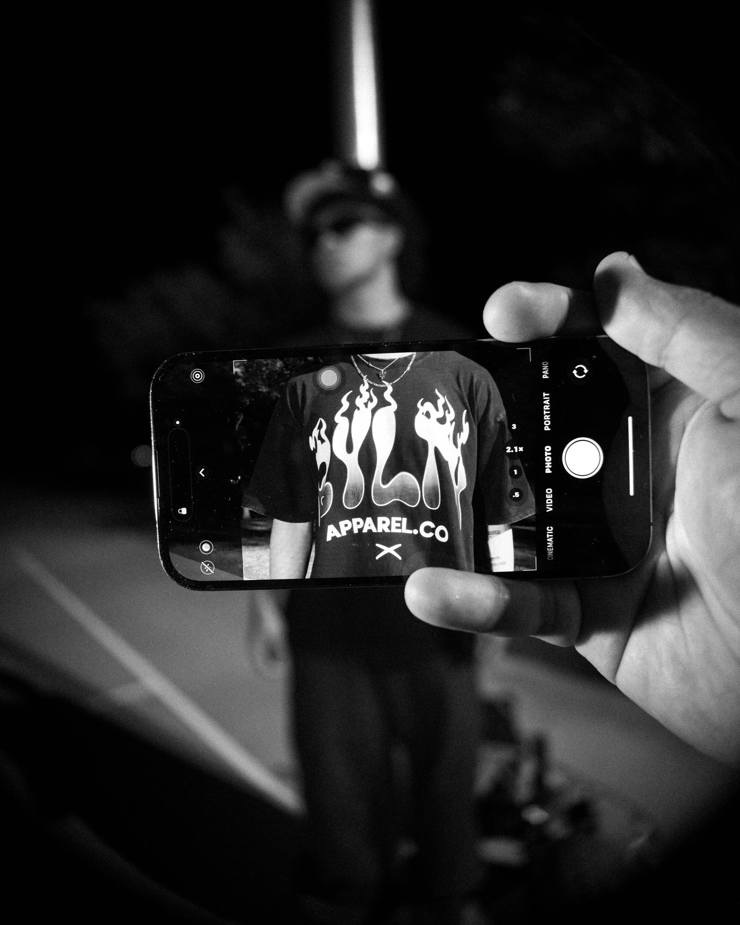 A person taking a selfie with a smartphone; the photo on the phone shows someone wearing a black t-shirt with flame graphics and the words 'APPAREL.CO'. The background is dark and blurry.