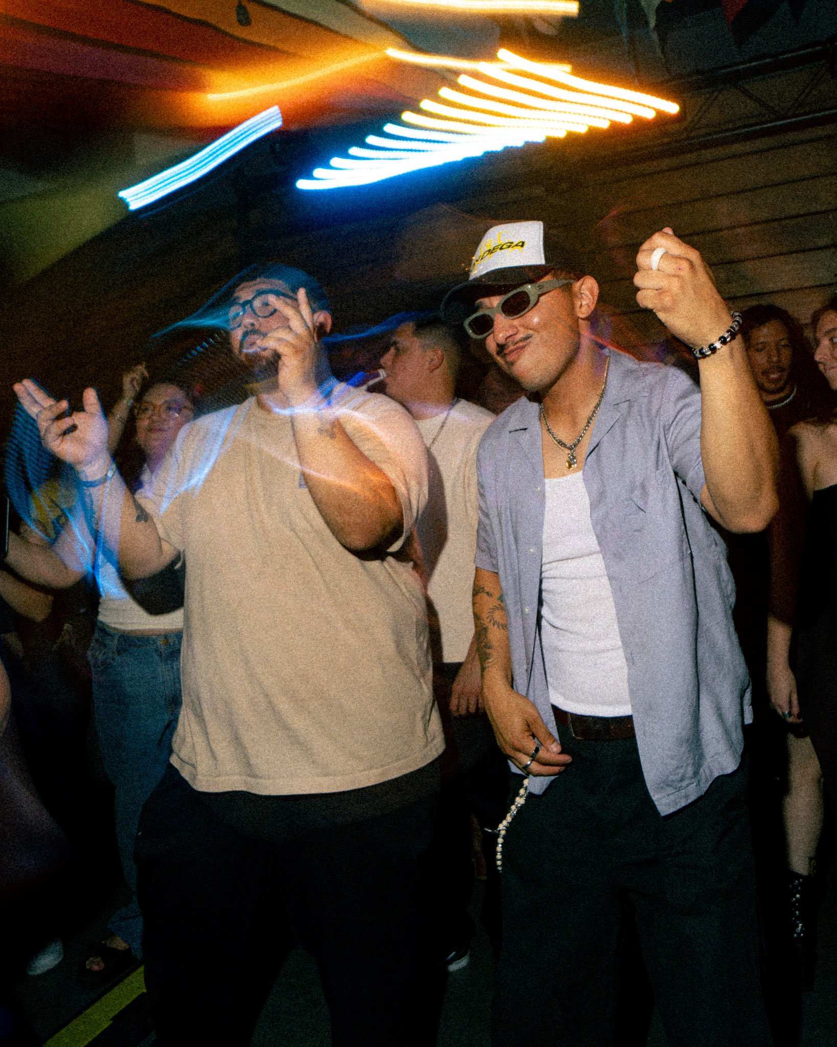 Two men are dancing and enjoying themselves at a nightclub or party under colorful lights. The man on the left wears glasses, a beige shirt, and has his eyes closed, while the man on the right wears sunglasses, a cap, and has tattoos on his arm. Both