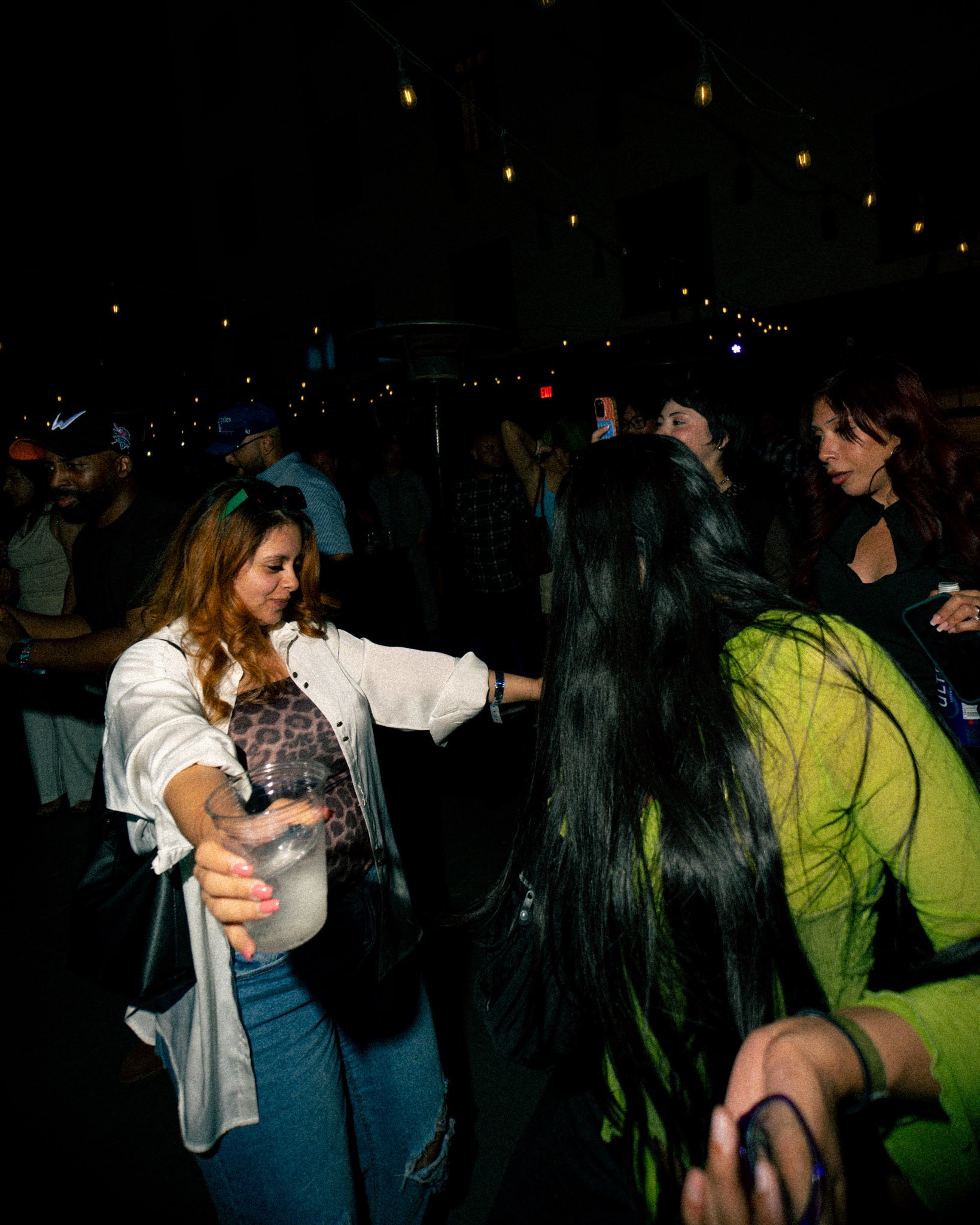 Group of people at a party or social event in a dimly lit room with string lights overhead, engaging in conversation.