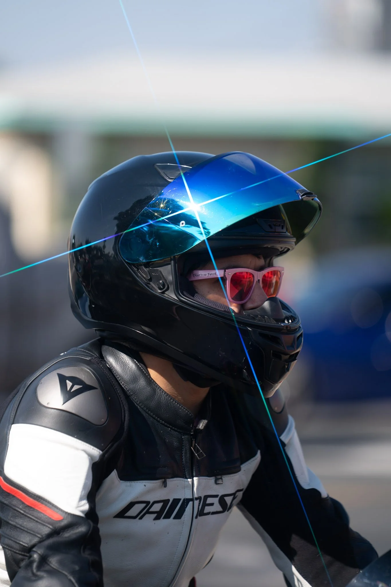 Person wearing a black full-face helmet, pink sunglasses, and a leather motorcycle jacket with blue, black, and white details.