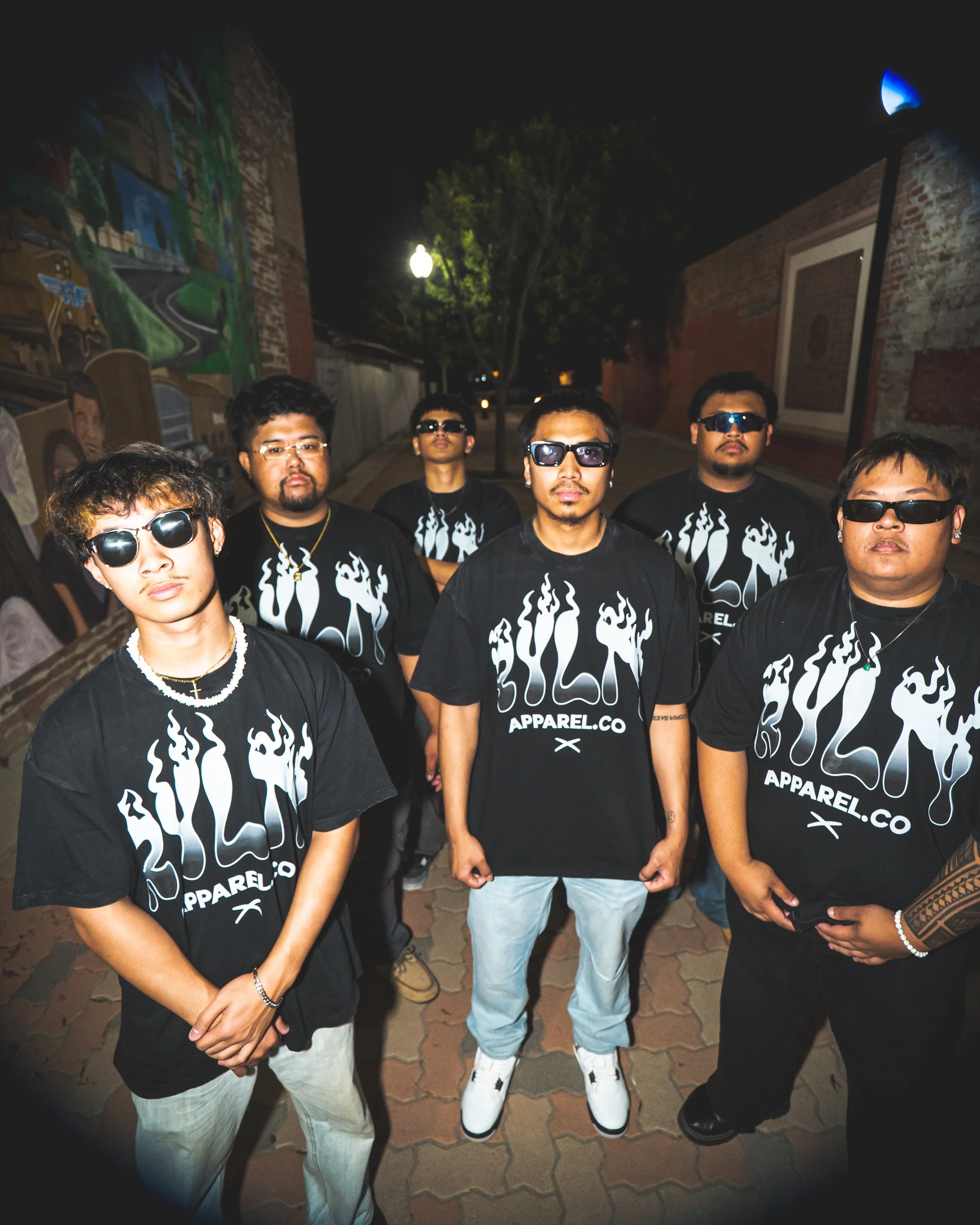 A group of six young men standing outdoors at night in a alleyway, wearing black T-shirts with white flame graphics and the words 'SOUL APPAREL CO'. Some are wearing sunglasses, and they have diverse hairstyles and facial hair. In the background, the
