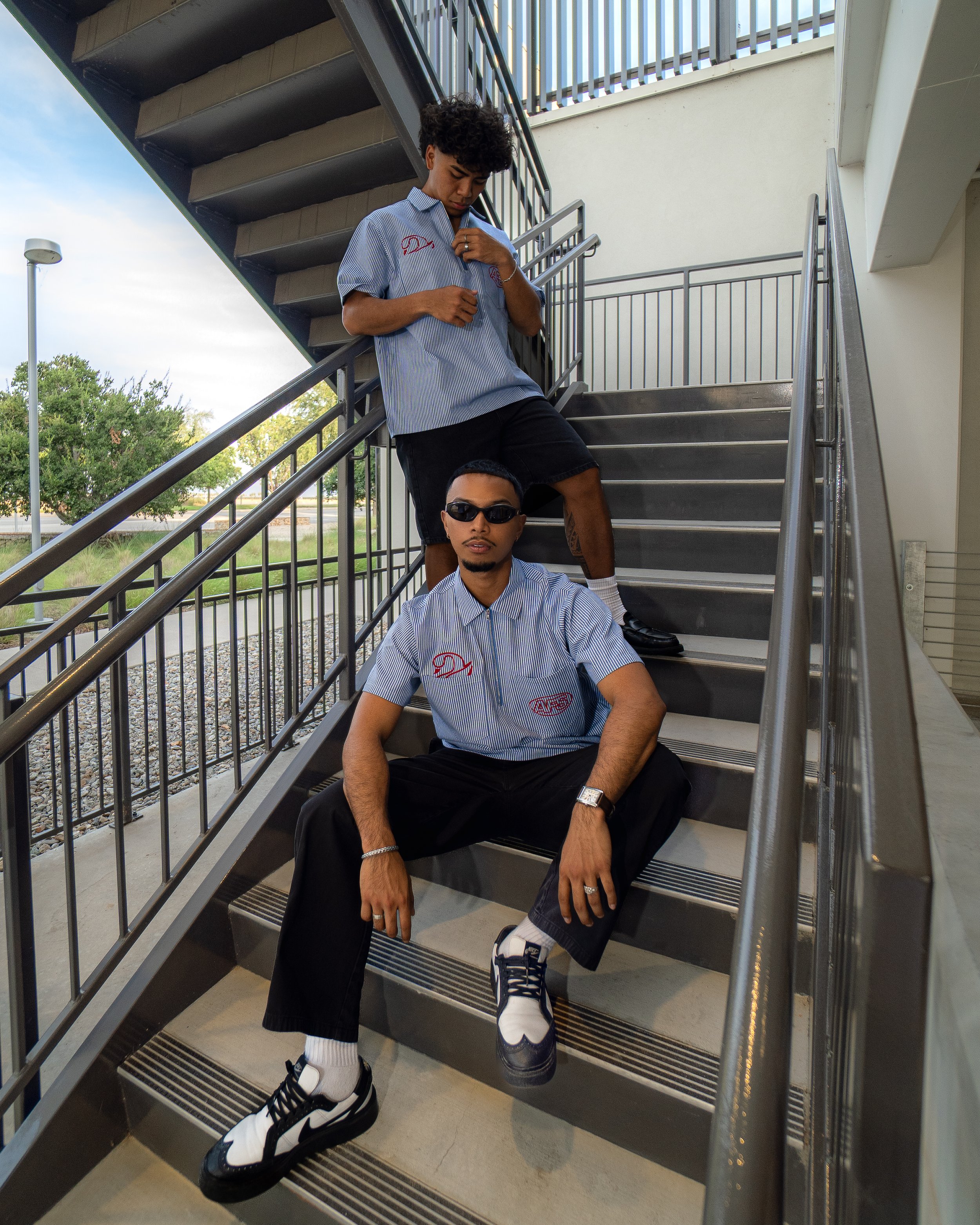 Three young men sitting and standing on an outdoor metal staircase with greenery and a pond visible in the background. They are dressed casually, with the two standing wearing matching light blue striped shirts with embroidered logos, and the seated 