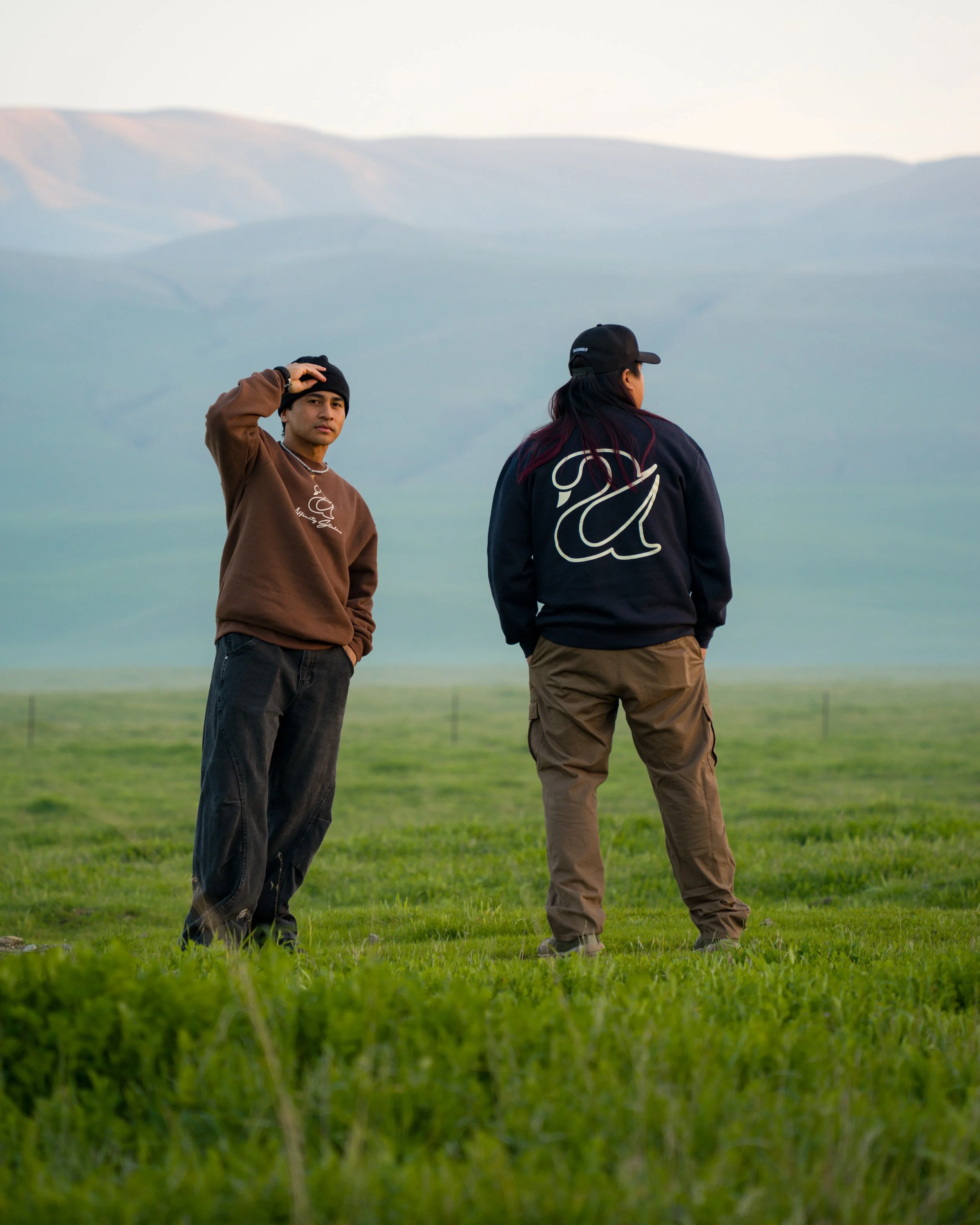 Two young adults, a man and a woman, standing in a lush green field with mountains in the background. The man is wearing a brown sweatshirt, black beanie, and has his hand on his head. The woman is dressed in a black jacket with a large white design,