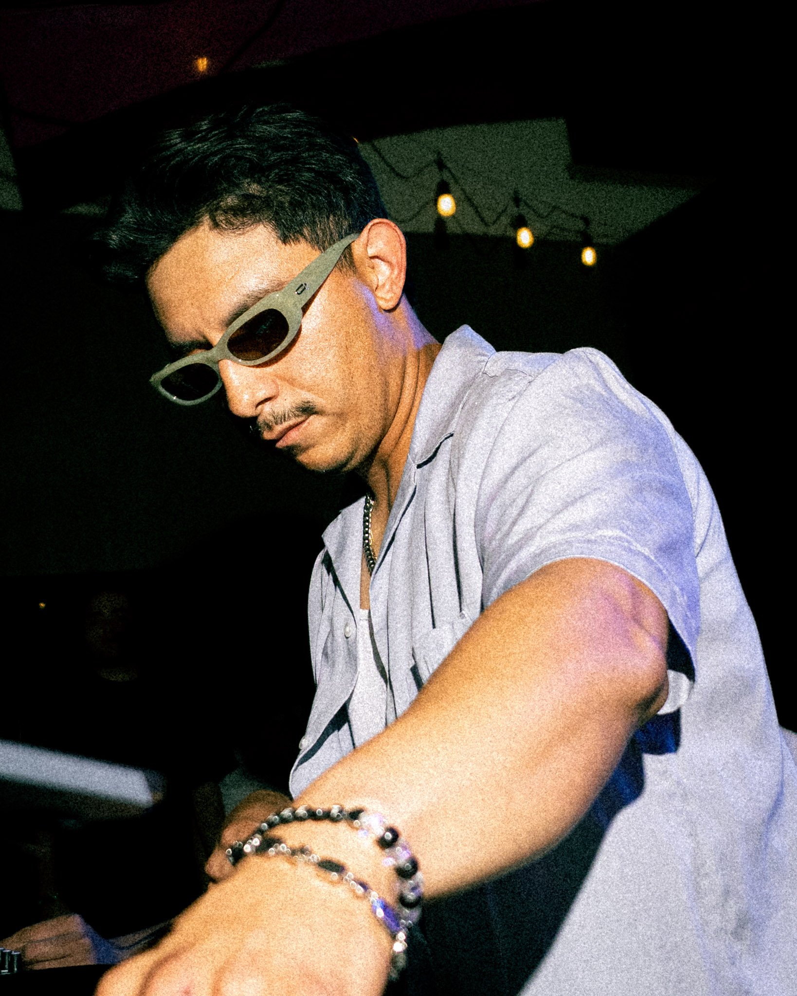 A young man with dark hair and glasses is wearing a light gray hoodie and several bracelets on his wrist. The background has dark lighting with some small, warm-colored lights hanging.