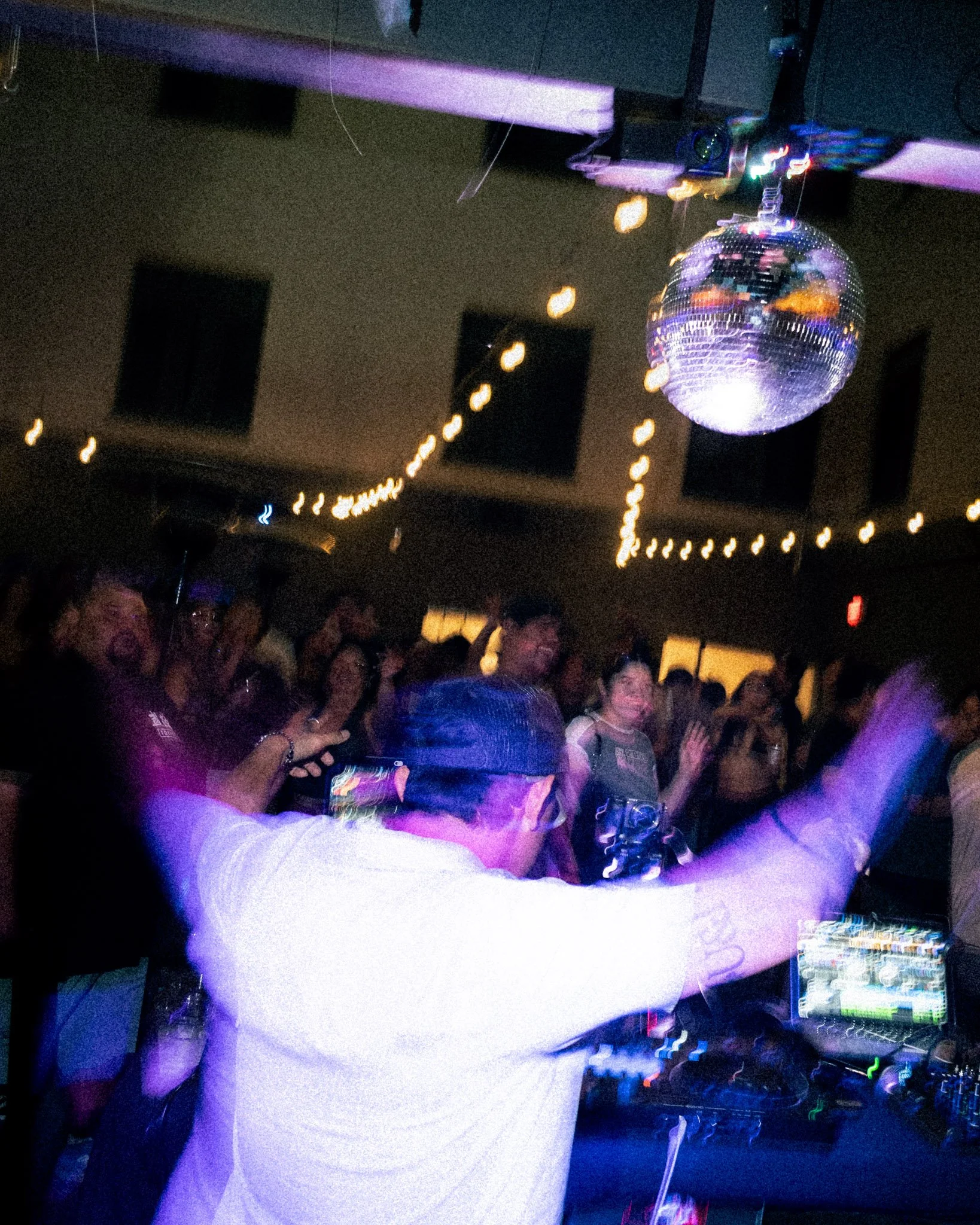 A man dressed in a white shirt is playing DJ at a nightclub, with a disco ball hanging from the ceiling reflecting colorful lights, and a crowd of people dancing and enjoying the music.