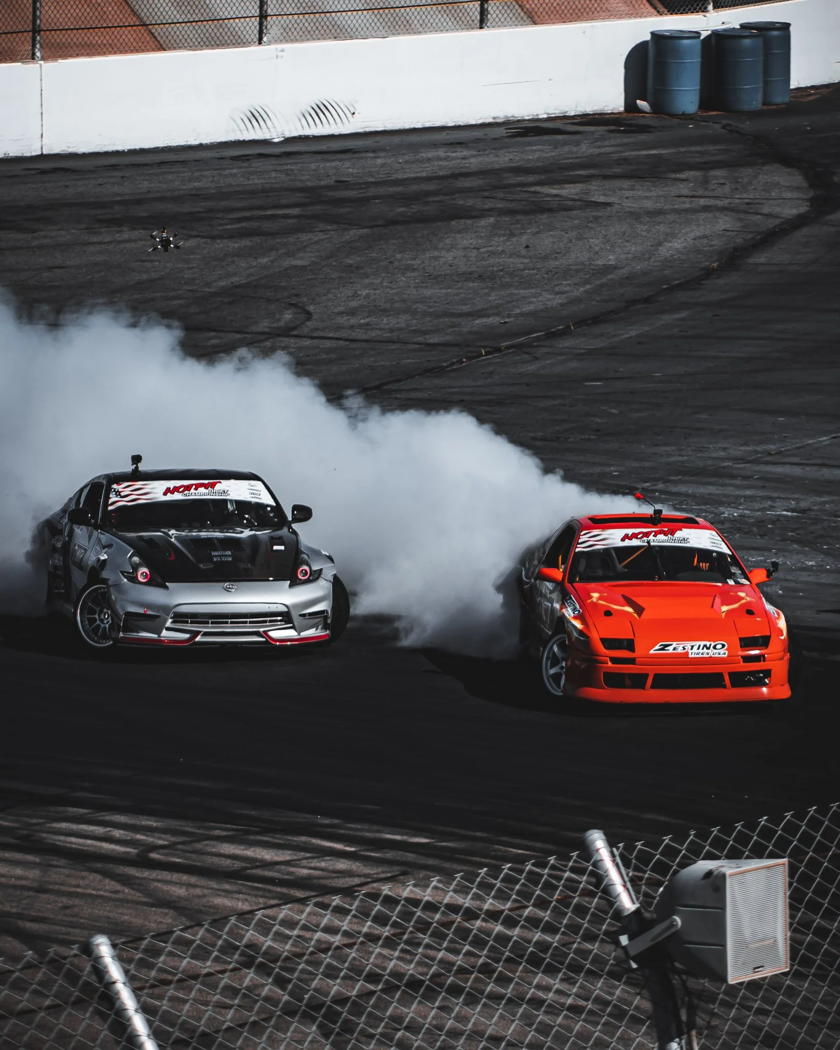 Two race cars drifting on a racetrack, creating smoke from their tires. The car on the left is gray with red accents, and the car on the right is orange with black accents. There is a small drone flying in the background near a white barrier with a drain, and three black trash barrels are placed against the barrier. The foreground is partially blocked by a chain-link fence.