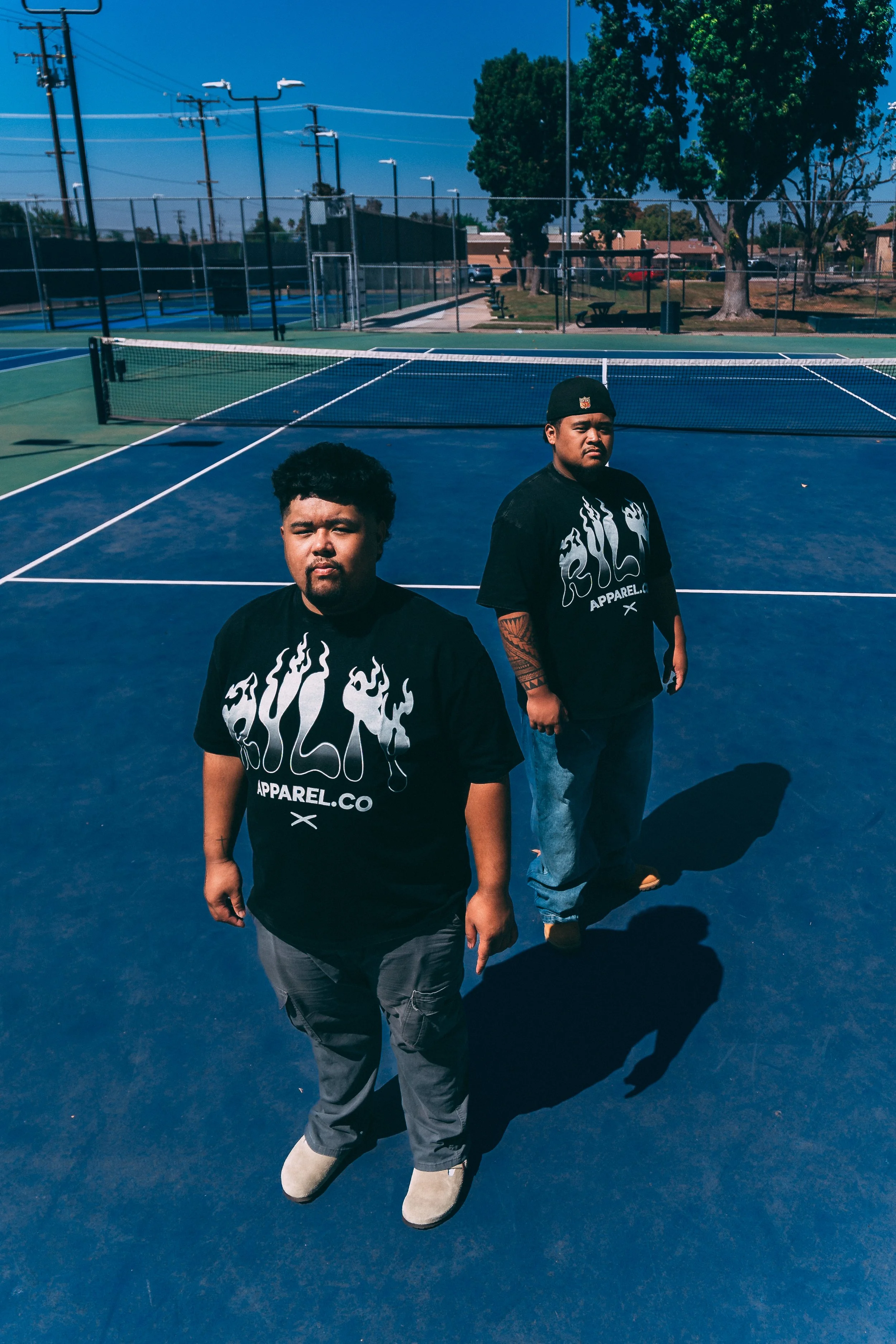 Two young men standing on a blue tennis court, with trees, a fence, and houses in the background. They are wearing black T-shirts with a white flame design and the word 'GUILT' and 'APPAREL.CO' printed on them.