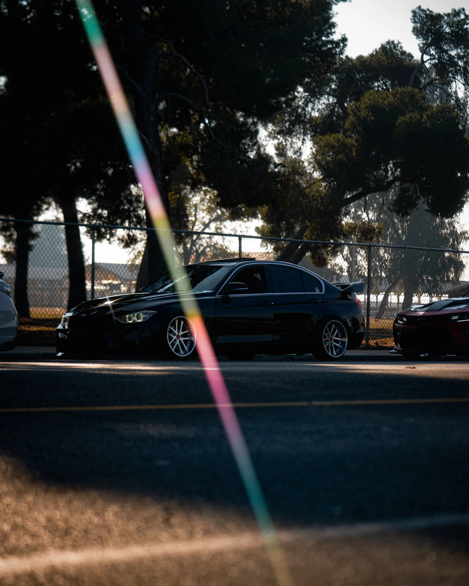 Black car parked near a chain-link fence with tall trees in the background, taken during late evening or early morning with sunlight casting shadows.