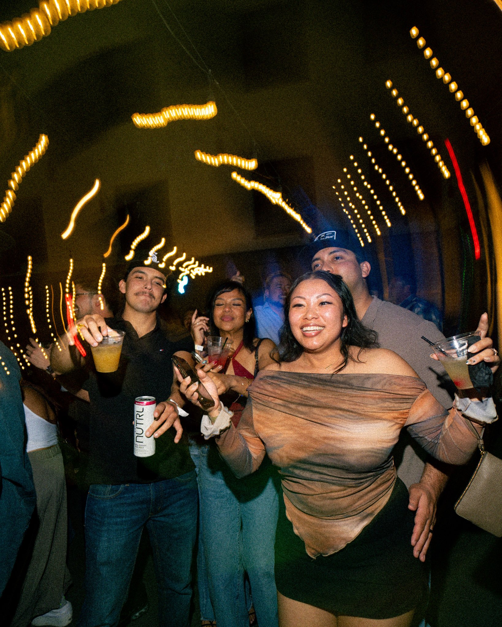 A group of people at a party holding drinks, with blurred, decorative string lights overhead creating motion effect.