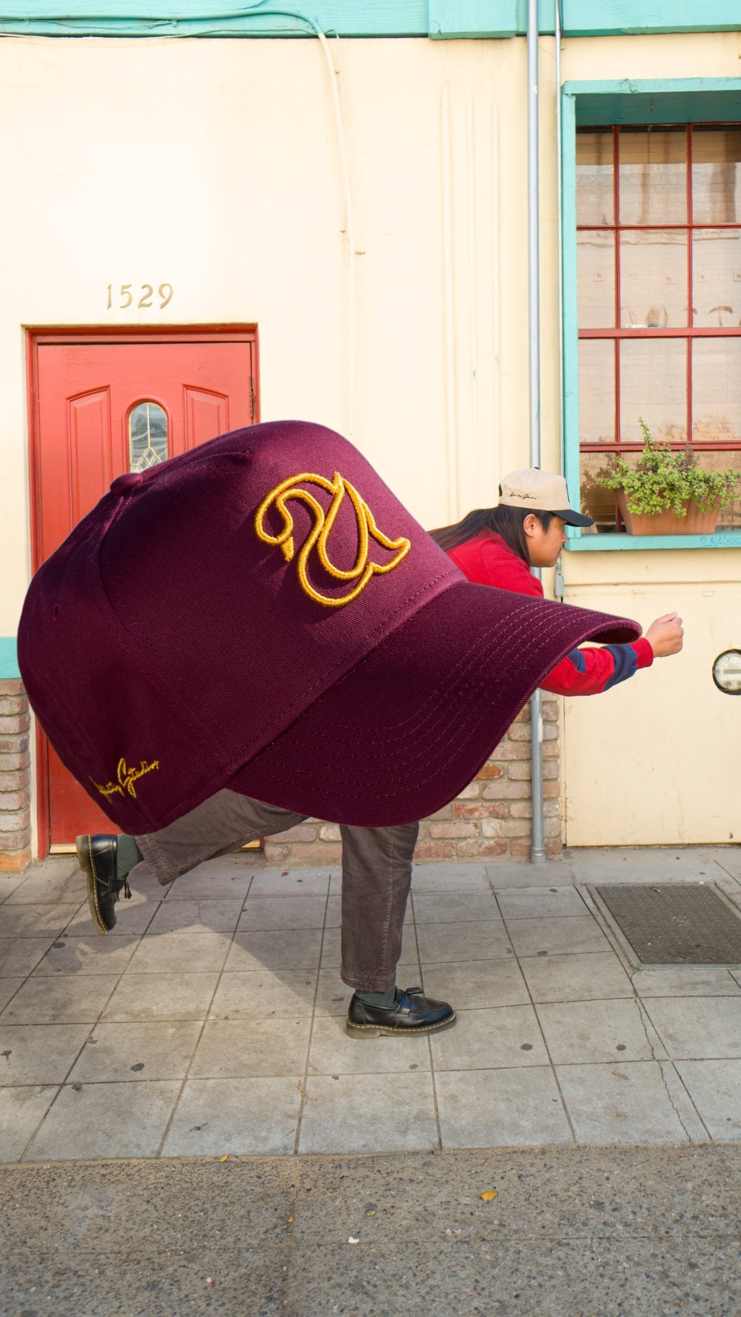 A person bending forward with their left leg raised, wearing a large maroon baseball cap with yellow embroidery, a red jacket, gray pants, and black shoes, standing on a sidewalk in front of a building with a red door and a window with a potted plant