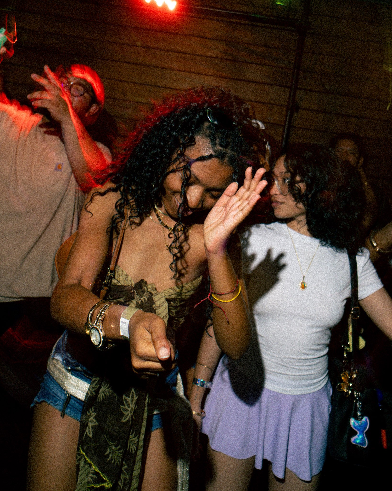 A group of young women at a party, with one wearing glasses and a beige top with a floral pattern, raising her hand to her mouth, another with curly hair dressed in a black and beige patterned top, and a third with dark hair and glasses wearing a whi