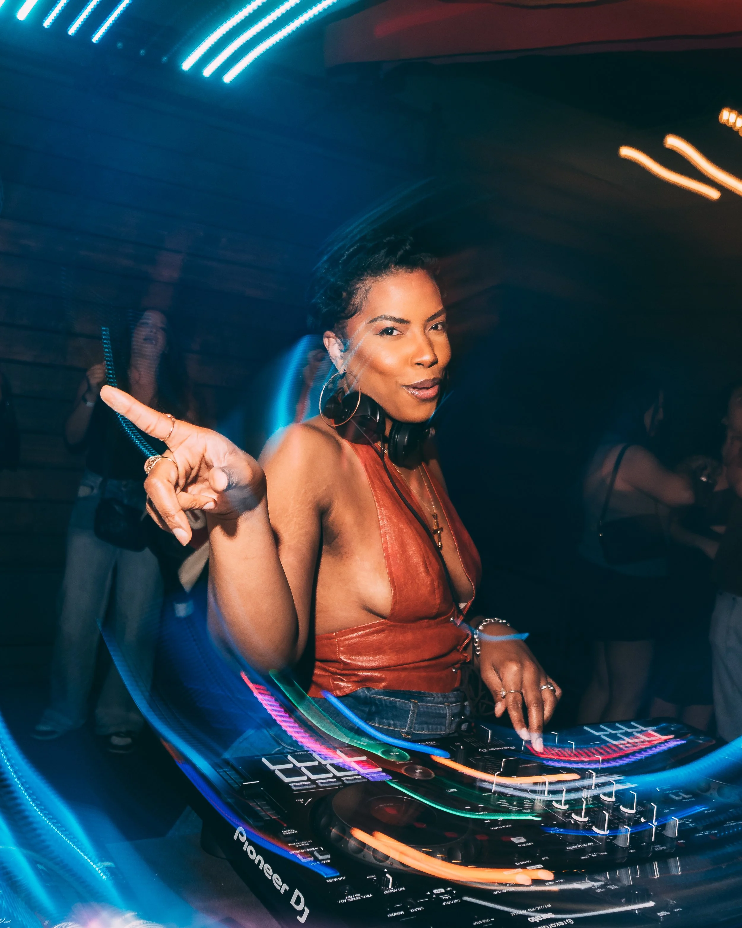 A woman with dark hair and hoop earrings smiling and pointing while DJing at a club or event with neon lights and a Pioneer DJ controller in front of her.