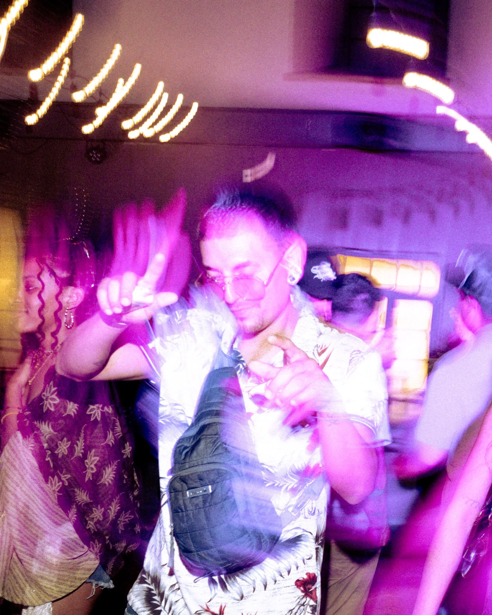 A woman wearing glasses and a floral dress dancing at a party with purple lighting.