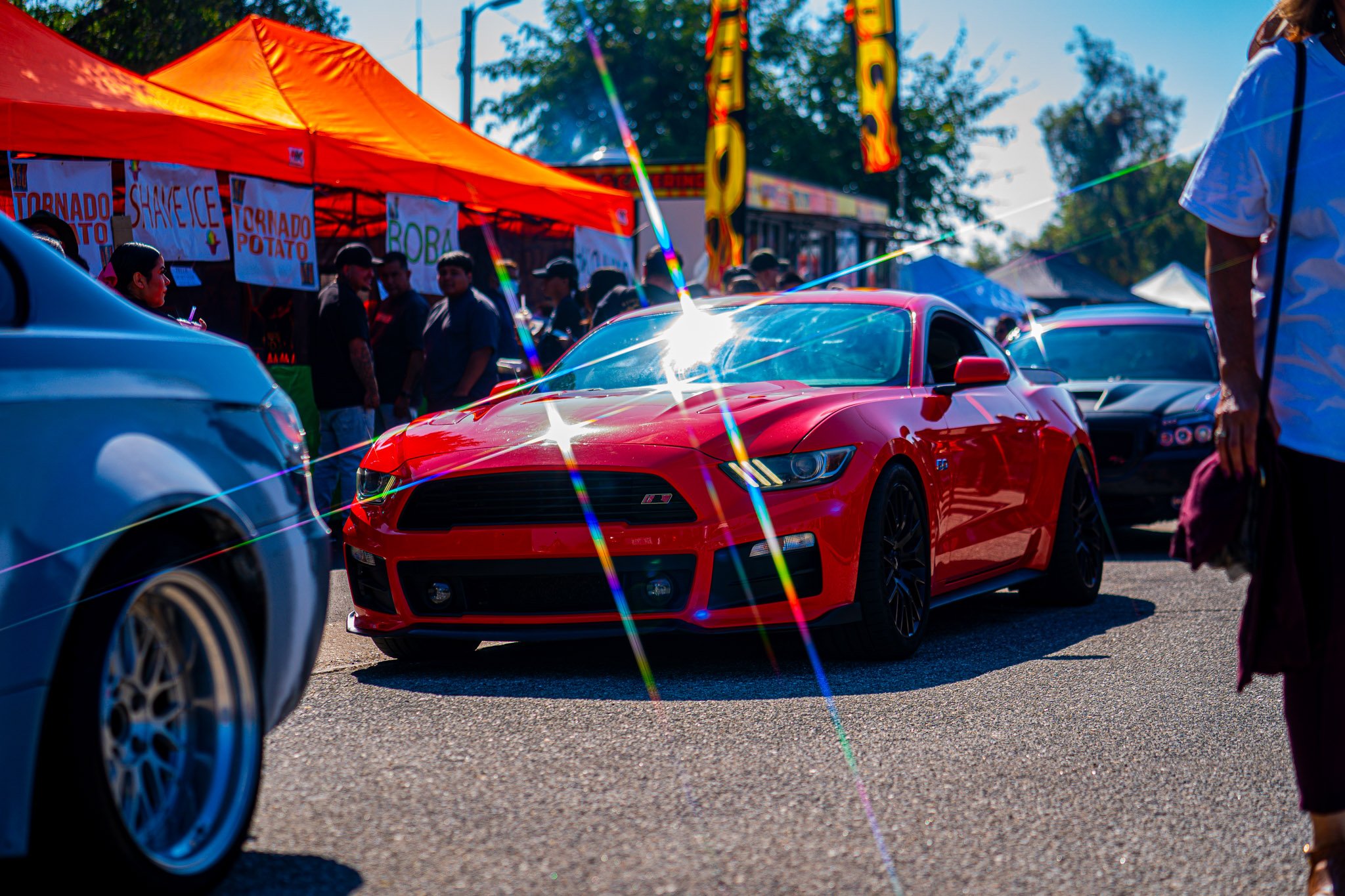 A red sports car parked at an outdoor event with an orange tent and a crowd of people in the background, and another vehicle partially visible to the side.