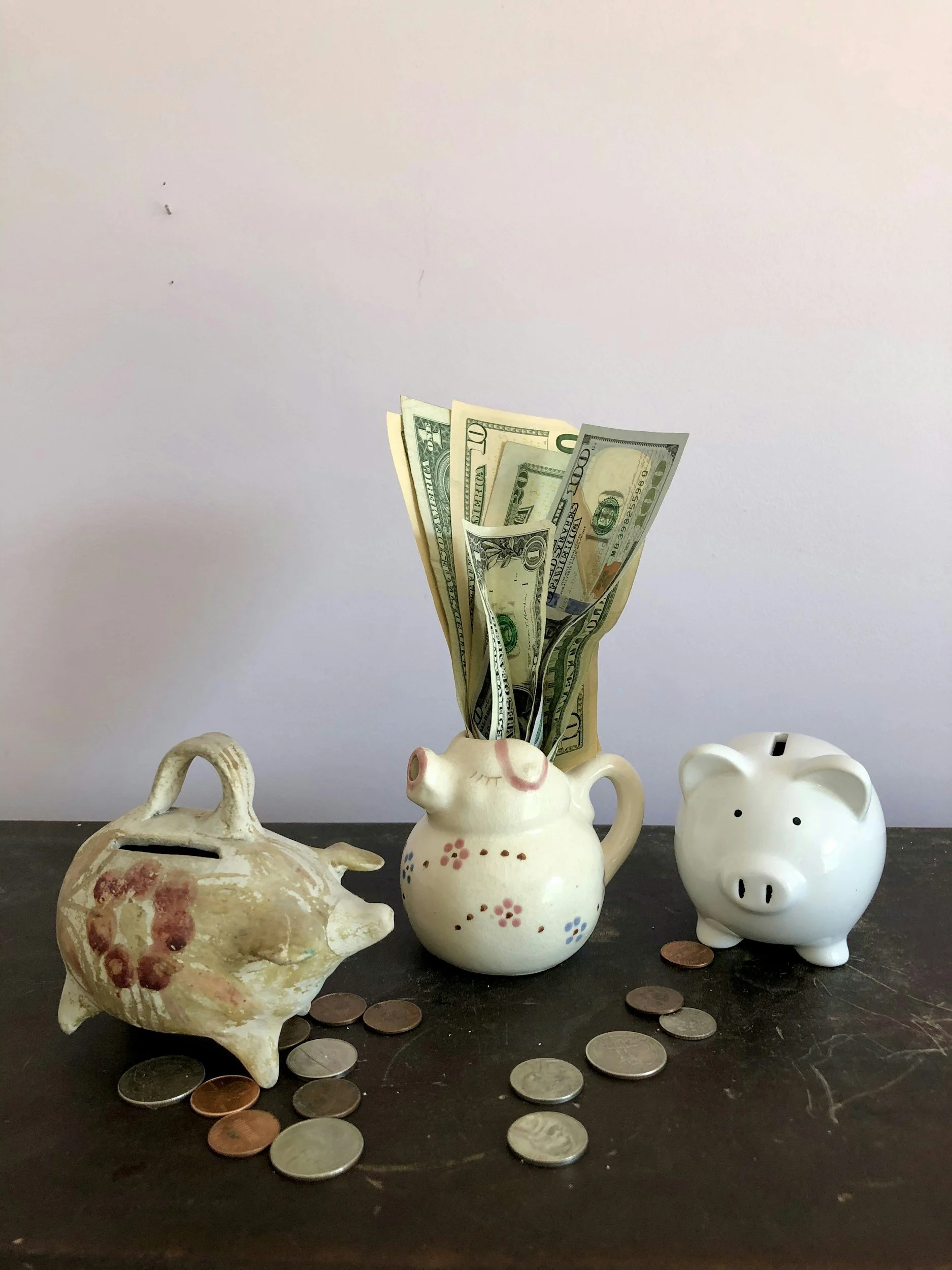 Three piggy banks: a vintage ceramic piggy bank on the left, a floral piggy bank with dollar bills in the middle, and a white ceramic piggy bank on the right, with scattered coins on a dark surface and a plain light-colored background.