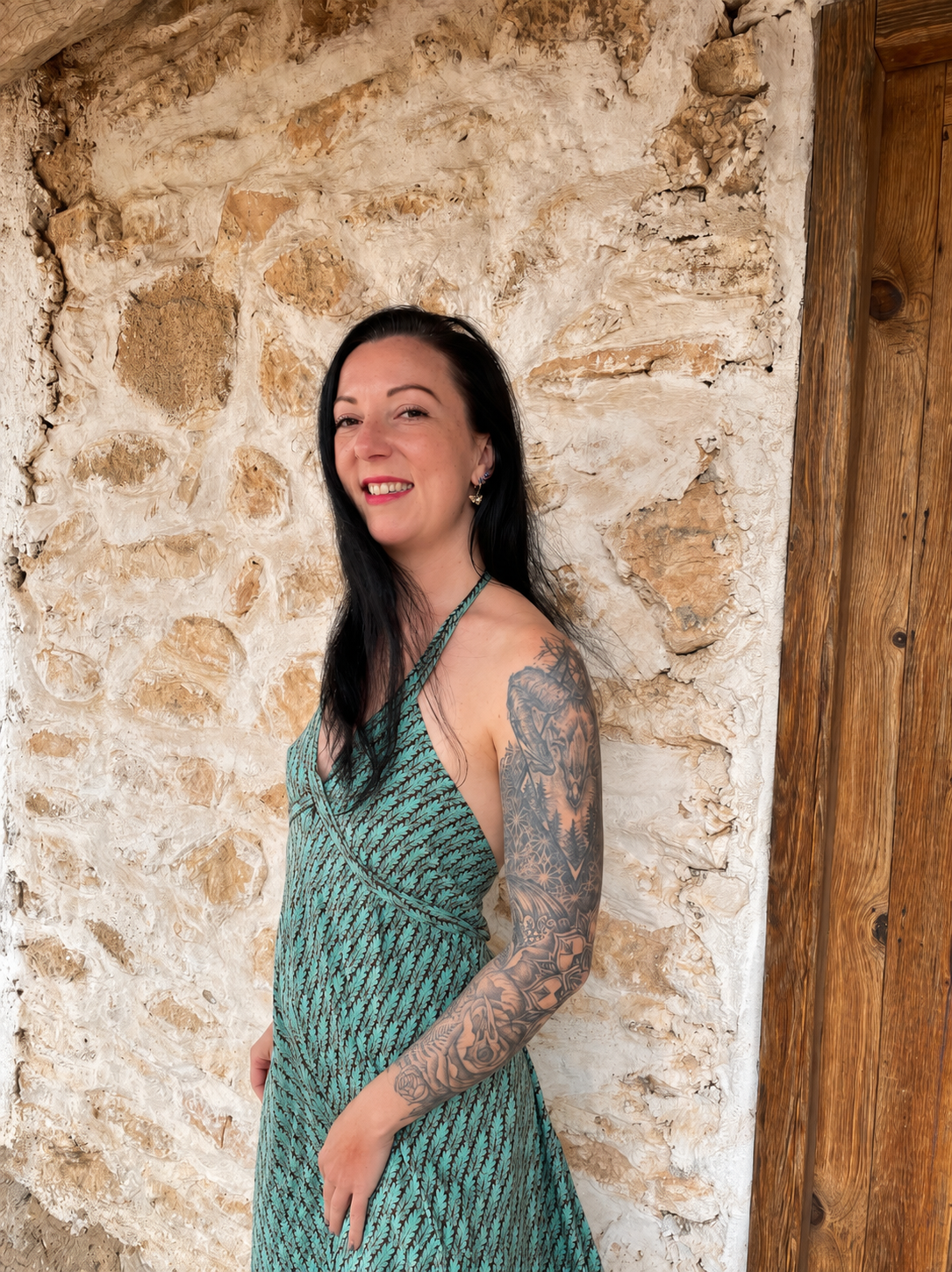 Junge Frau mit schwarzen Haaren, rotem Lippenstift und Tattoo-Arm, trägt ein grünes Kleid, steht vor einer alten Steinmauer mit Holzrahmen.