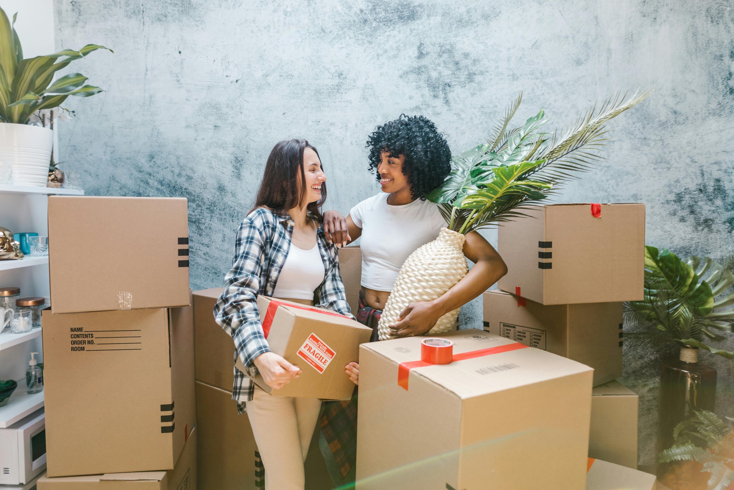 Two women smiling at each other among moving boxes, one holding a small boxed item and the other holding a large woven vase with green foliage, in a room with a textured wall and shelves with various items.
