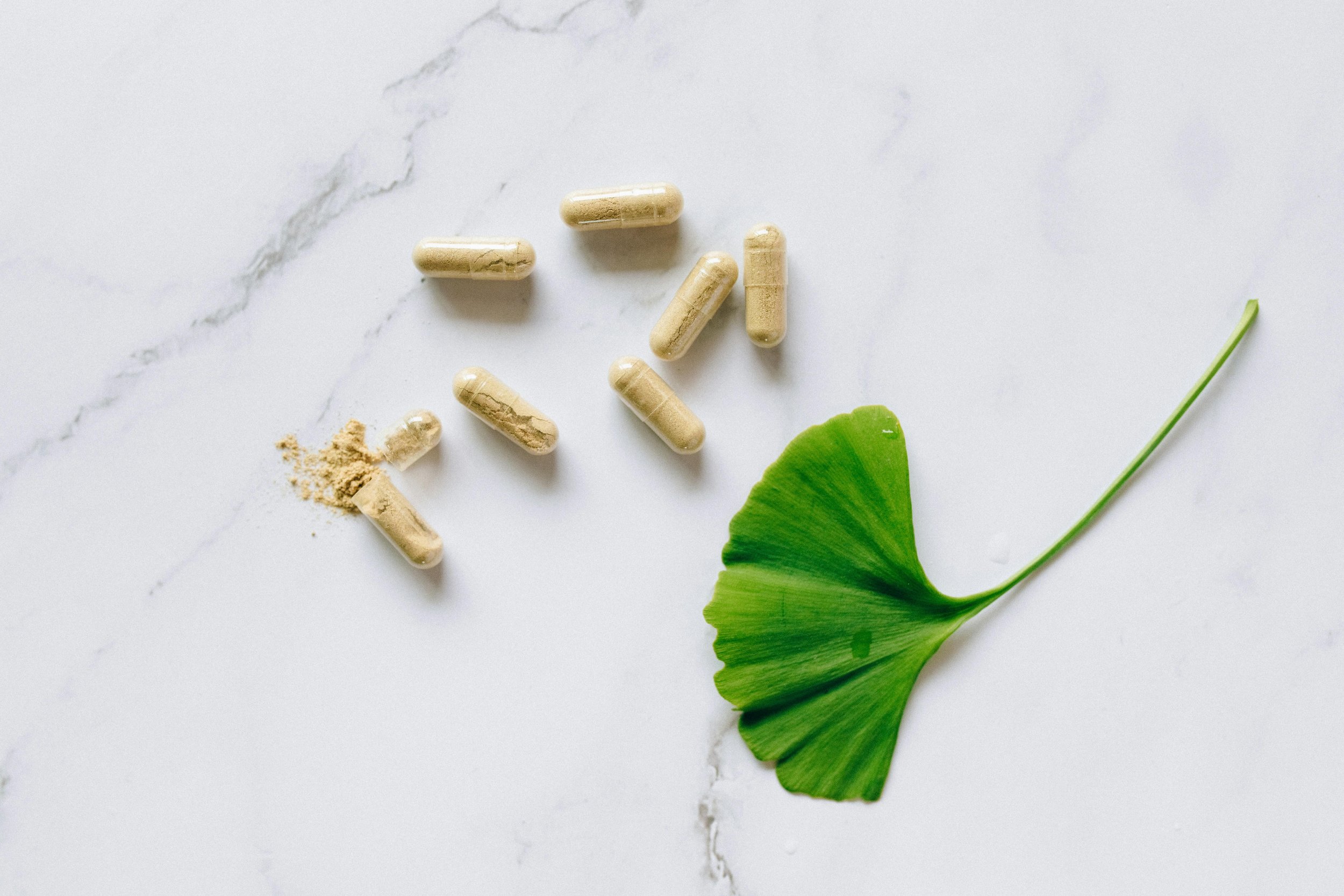 Several beige capsules, some broken open with powder spilled, are scattered on a white marble surface. A green ginkgo leaf is in the lower right corner.