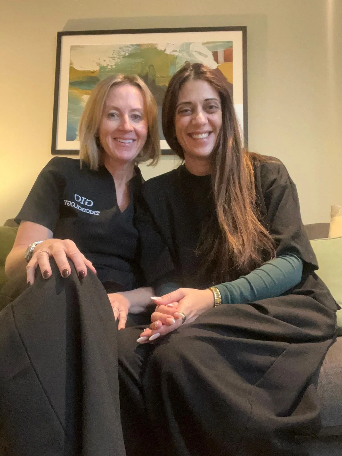 Two women sitting close together, smiling at the camera in a cozy room with a framed landscape painting behind them. One woman has blonde hair, wearing black scrubs with a logo, and the other has long brown hair, also dressed in black.