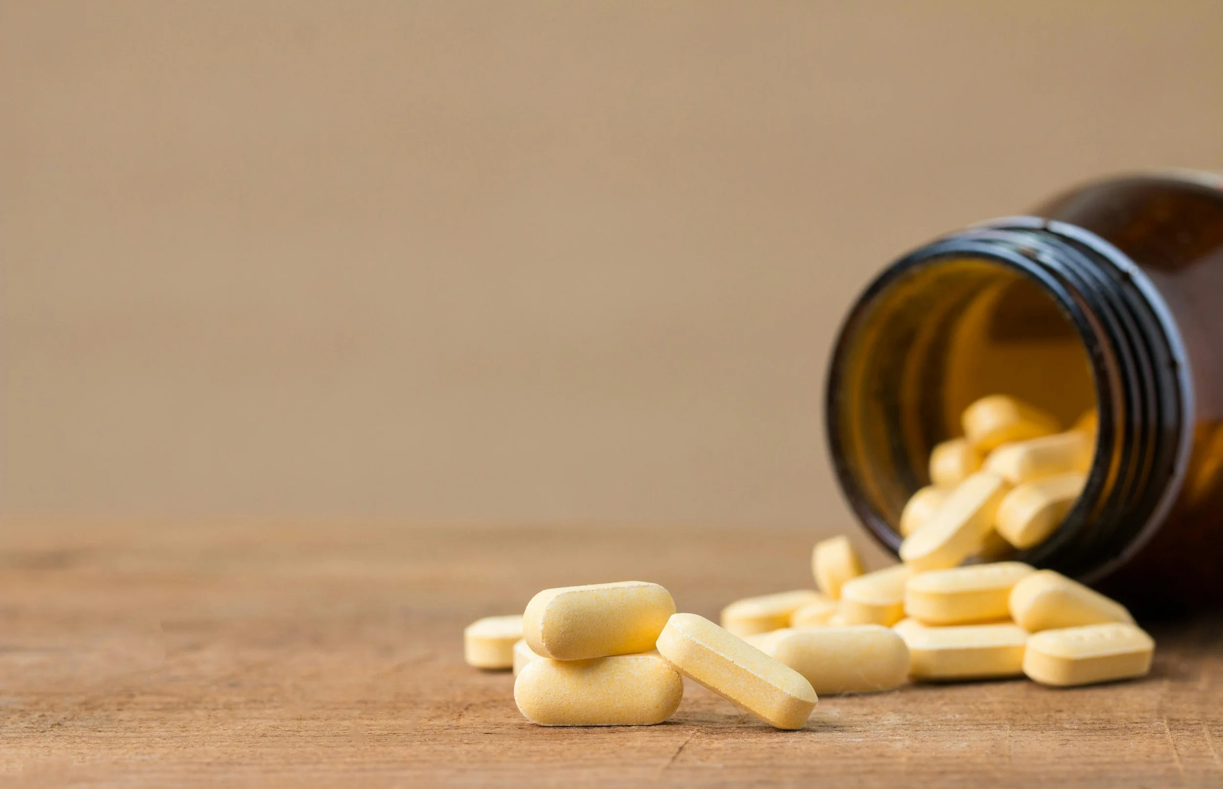 A brown prescription pill bottle laying on its side on a wooden surface, with yellow oval pills spilling out of it.