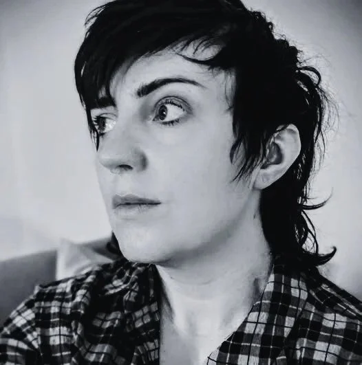 Black-and-white portrait of a woman with short, dark, messy hair, wearing a plaid shirt, looking to the side.