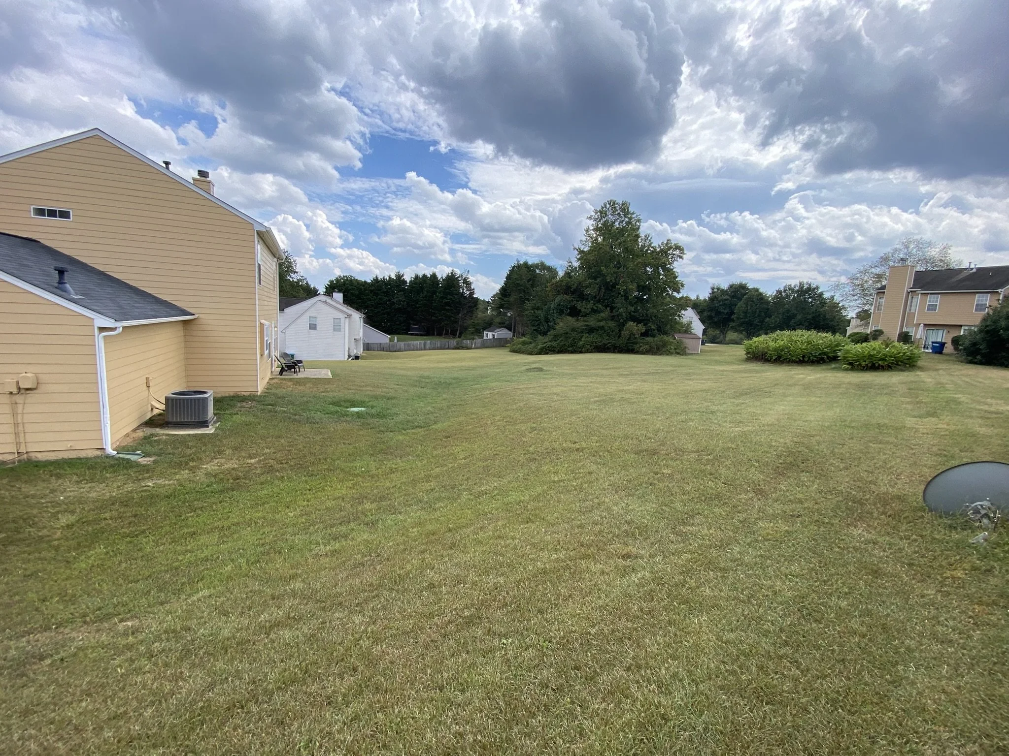 A wide grassy backyard with houses on each side, bushes, trees, and a cloudy sky.