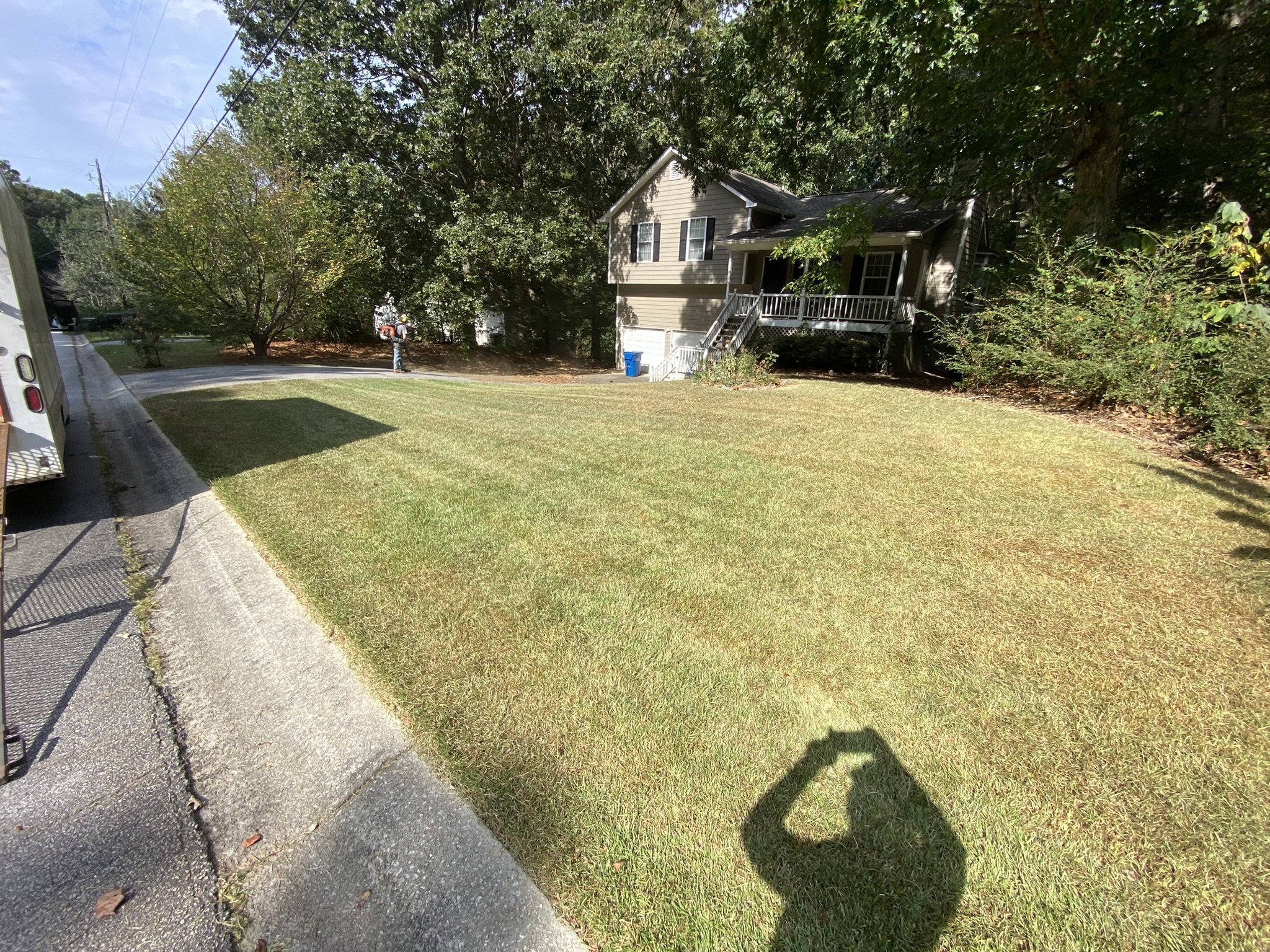 A residential yard with a well-maintained lawn, trees, and bushes. There is a person in the distance near the trees, and a house with a porch and stairs. A shadow of the person taking the photo is visible on the grass, and part of a trailer or vehicle is on the left.