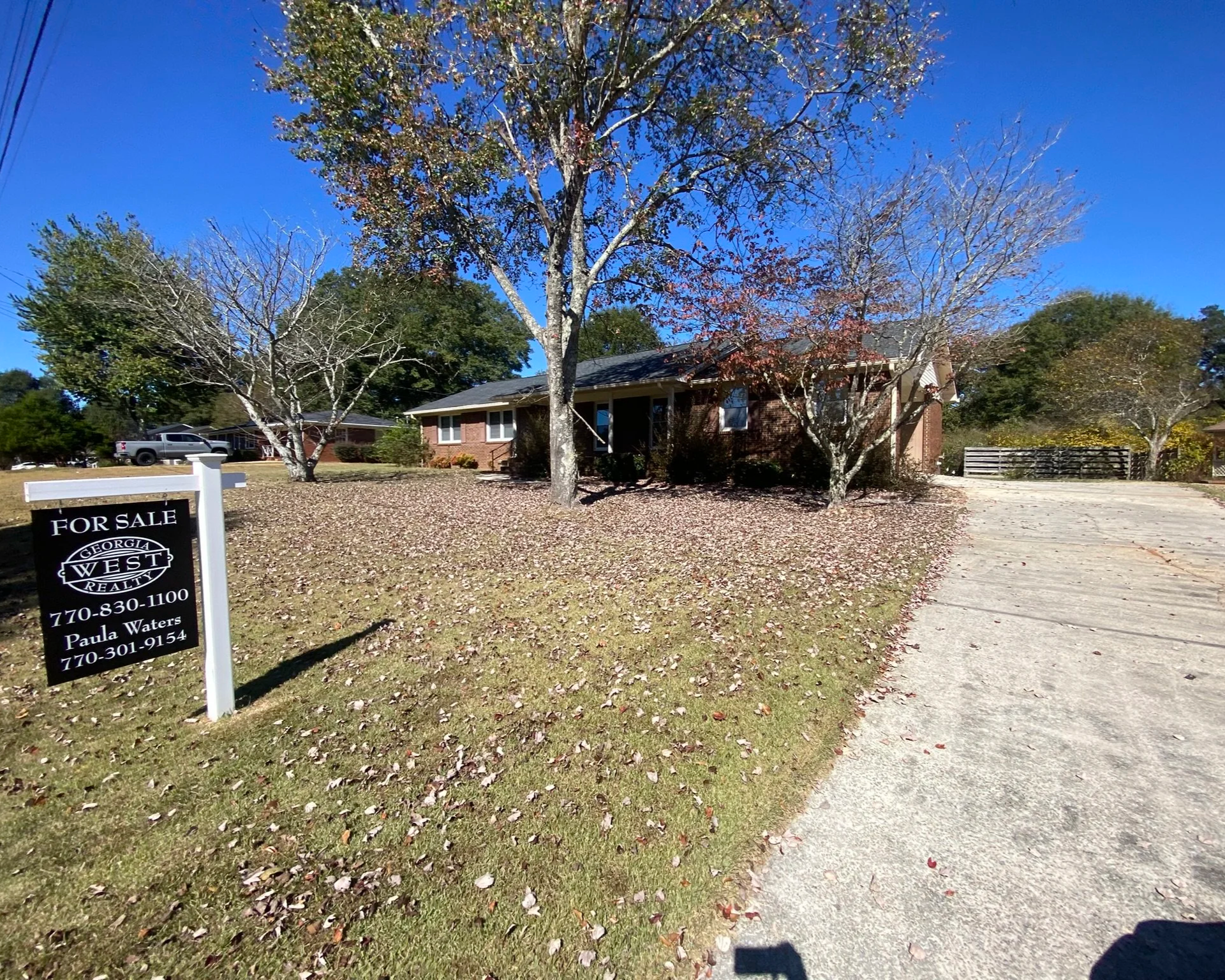 A brick house with a front yard and trees, a driveway on the right side, and a sign that says 'For Sale' by Georgia West Realty with contact information.