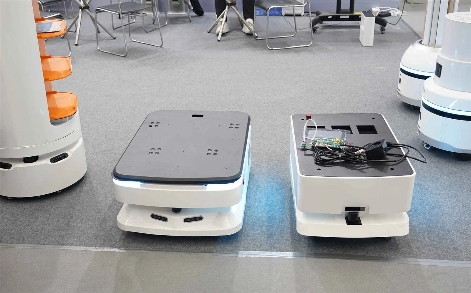 Two autonomous delivery robots on a gray carpeted floor in an indoor setting, with chairs and tables visible in the background.