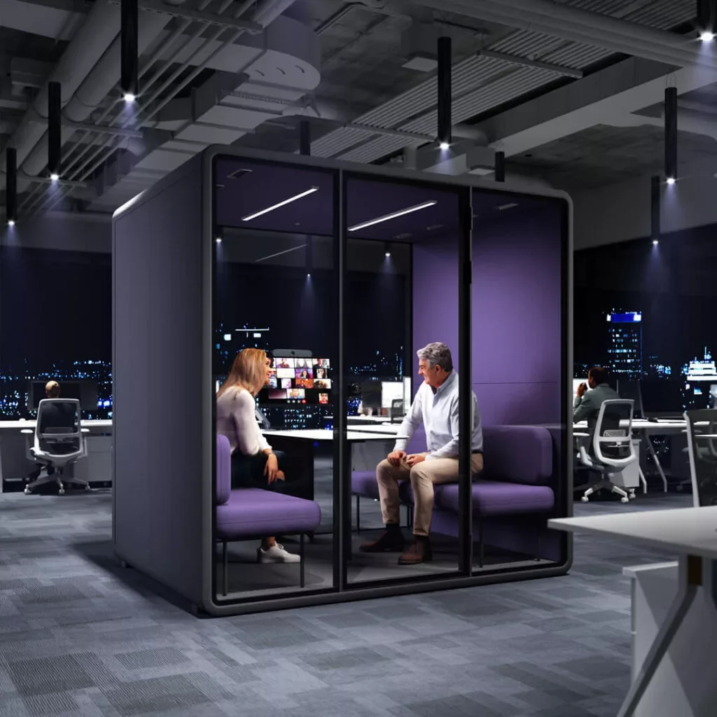 Two people are sitting inside a glass-walled phone booth in a modern office at night, having a conversation. The office has high ceilings, cityscape views through large windows, and several employees working at desks with computers.