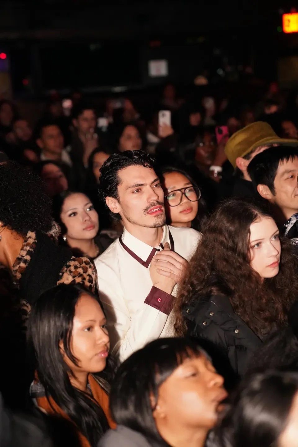 Crowd of diverse people at an indoor event, some with focused expressions, in dim lighting. Women's Magazine