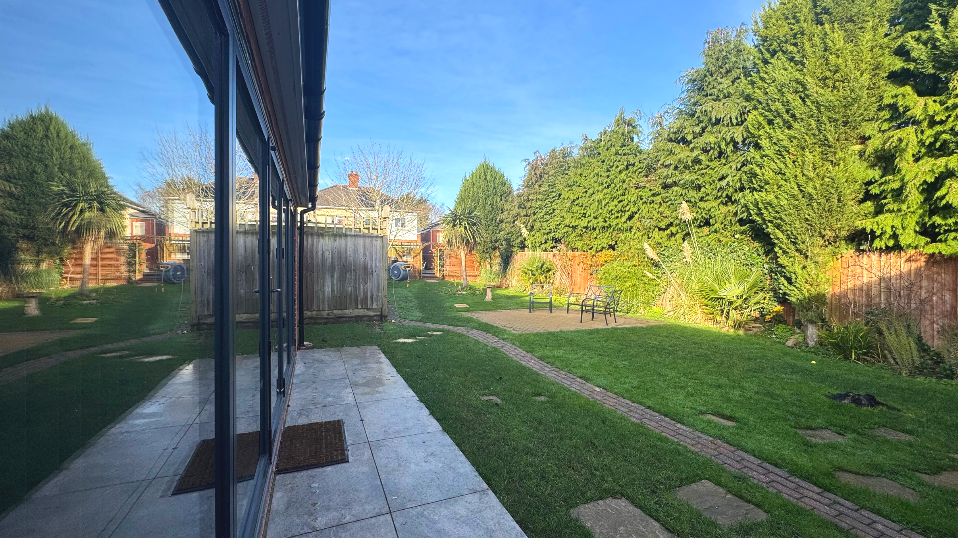 A backyard with a glass sliding door patio, green grass, a brick pathway, bushes, trees, and benches.