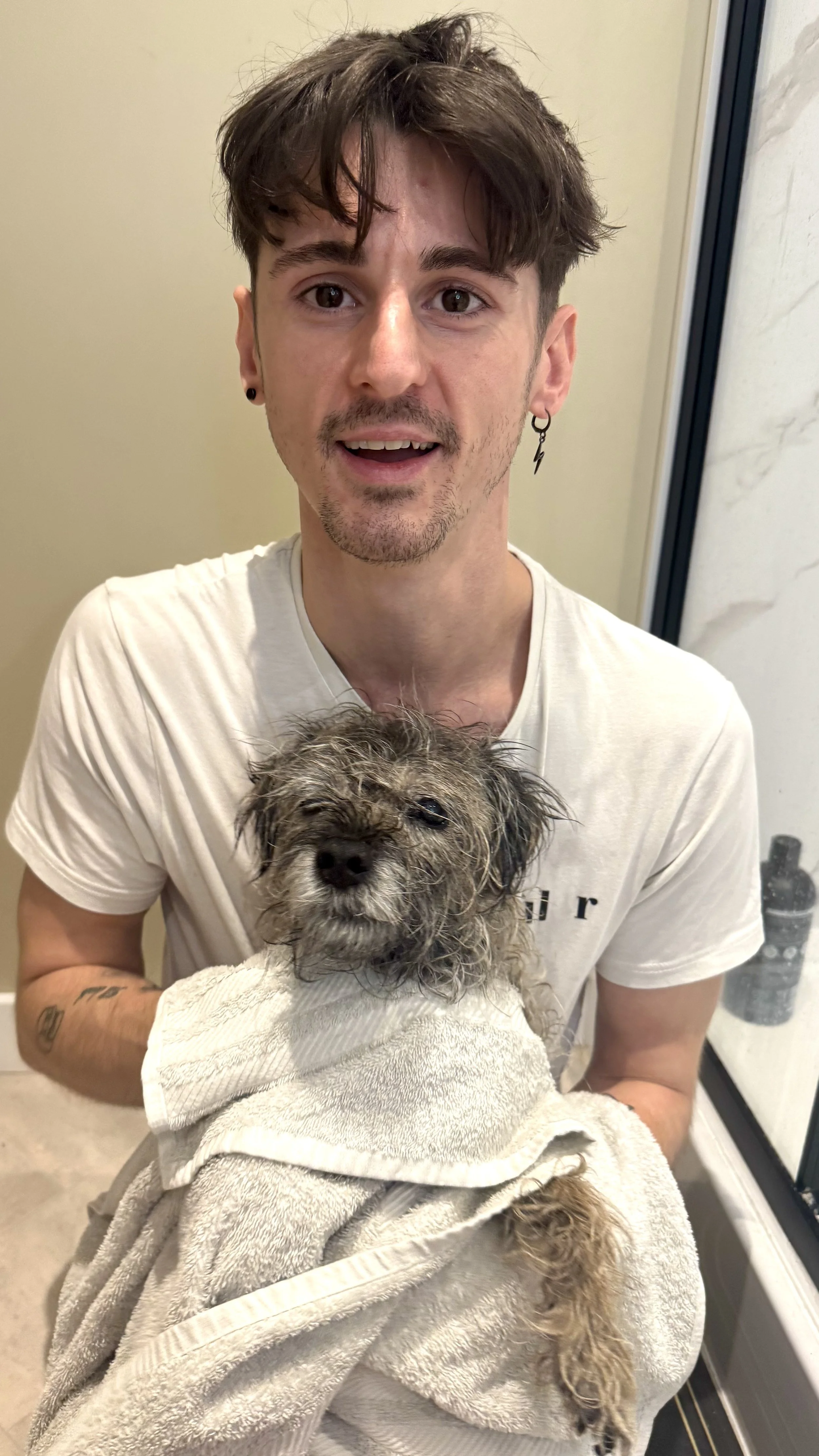 A man with brown hair, wearing earrings, holding a wet dog wrapped in a towel.