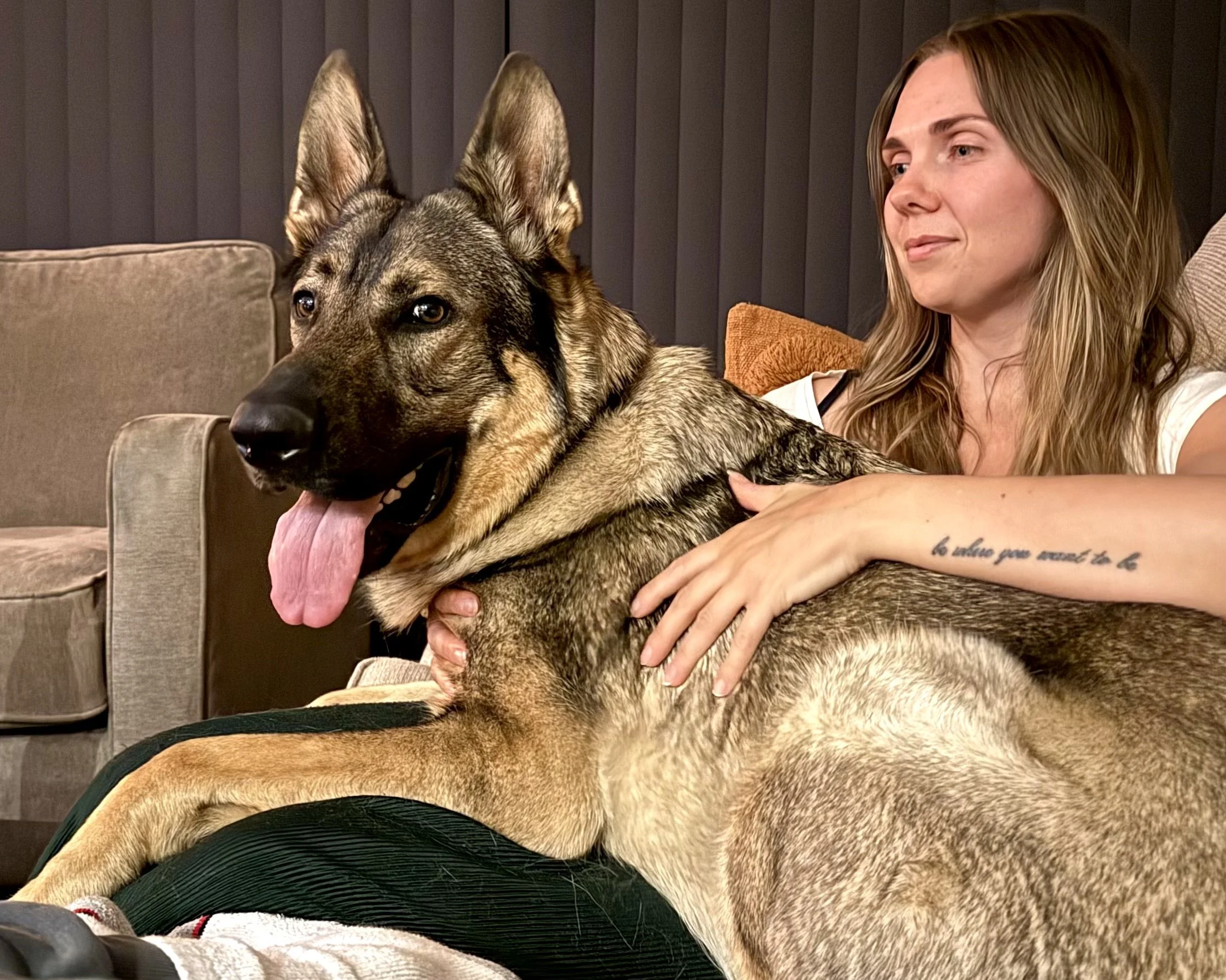 A woman sitting on a couch with a large German Shepherd dog resting on her lap, she is holding the dog gently and appears to be smiling softly, with a dark curtain and a beige couch in the background.