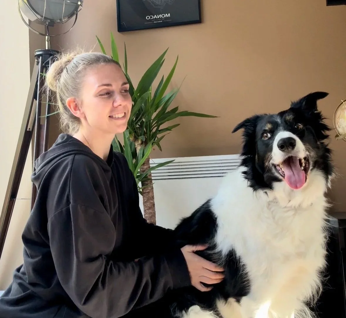 A woman with blonde hair in a bun smiling and holding a black and white Border Collie dog indoors, with a large green plant behind them.