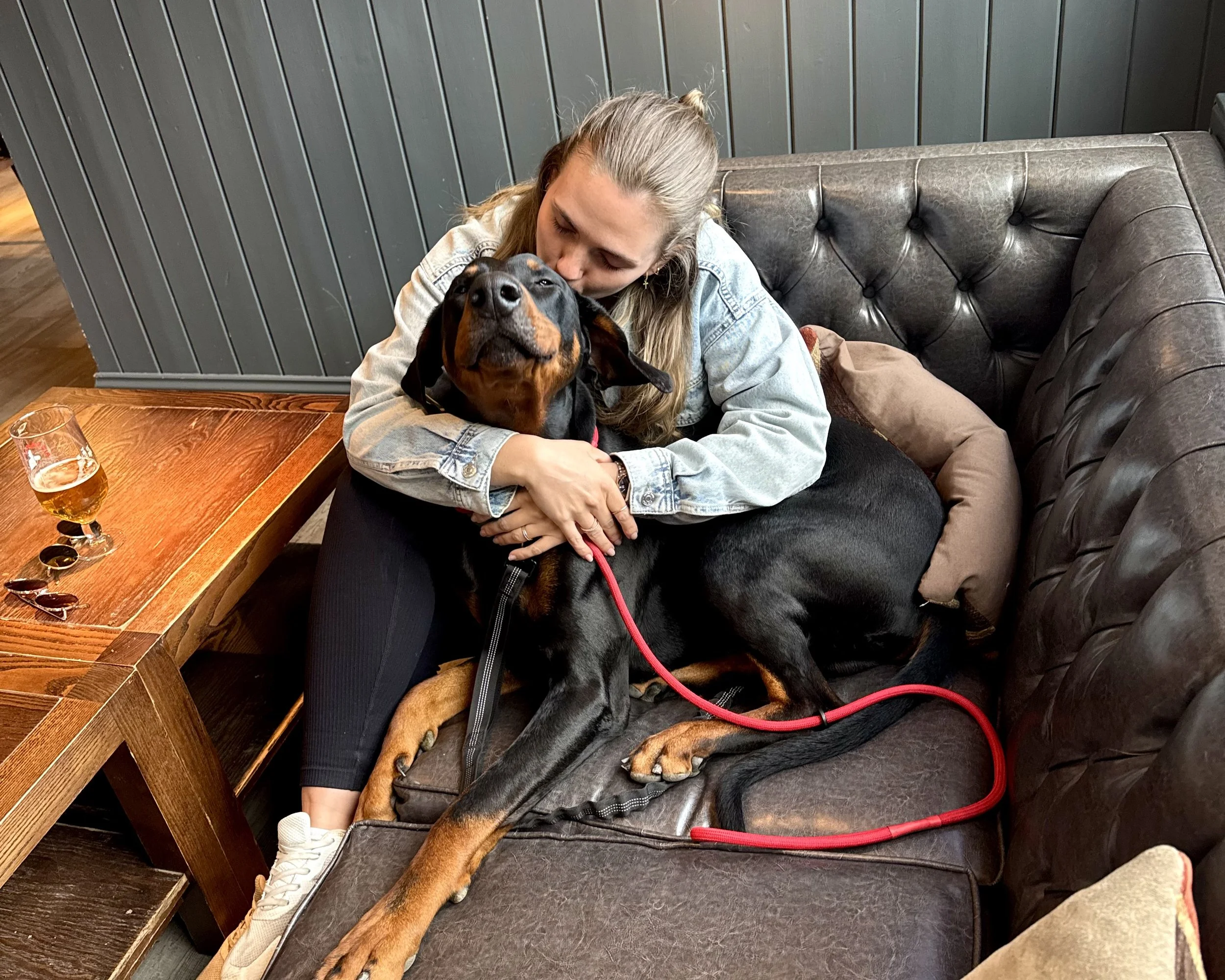 A woman with long blonde hair wearing a denim jacket hugging a large black and tan dog on a leather couch in a cozy cafe.