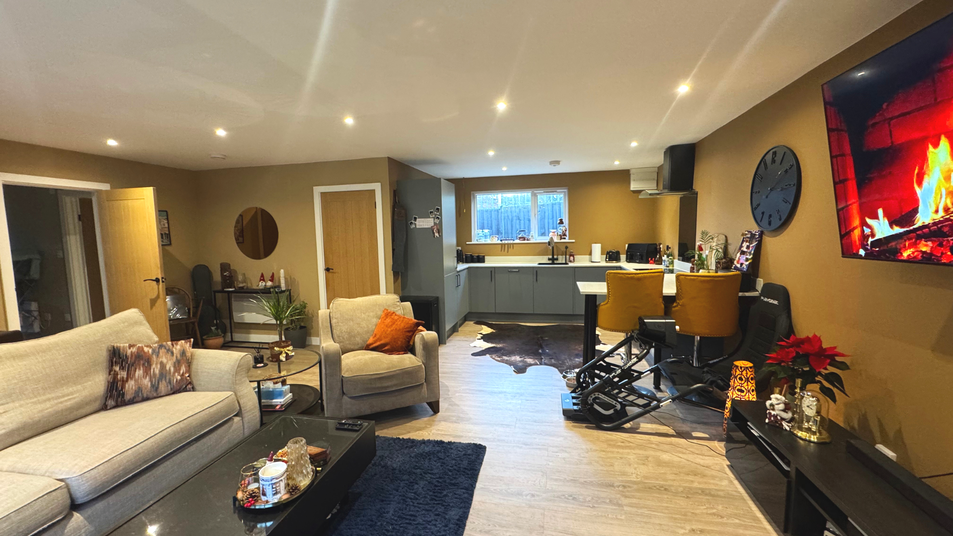 A spacious living room with beige furniture, a black coffee table, and a blue rug. The space includes a kitchen with gray cabinets, a window, and various appliances. There is a wall clock and a TV displaying a fireplace scene. Christmas decorations are seen on a black console table, and a treadmill is positioned near the wall.