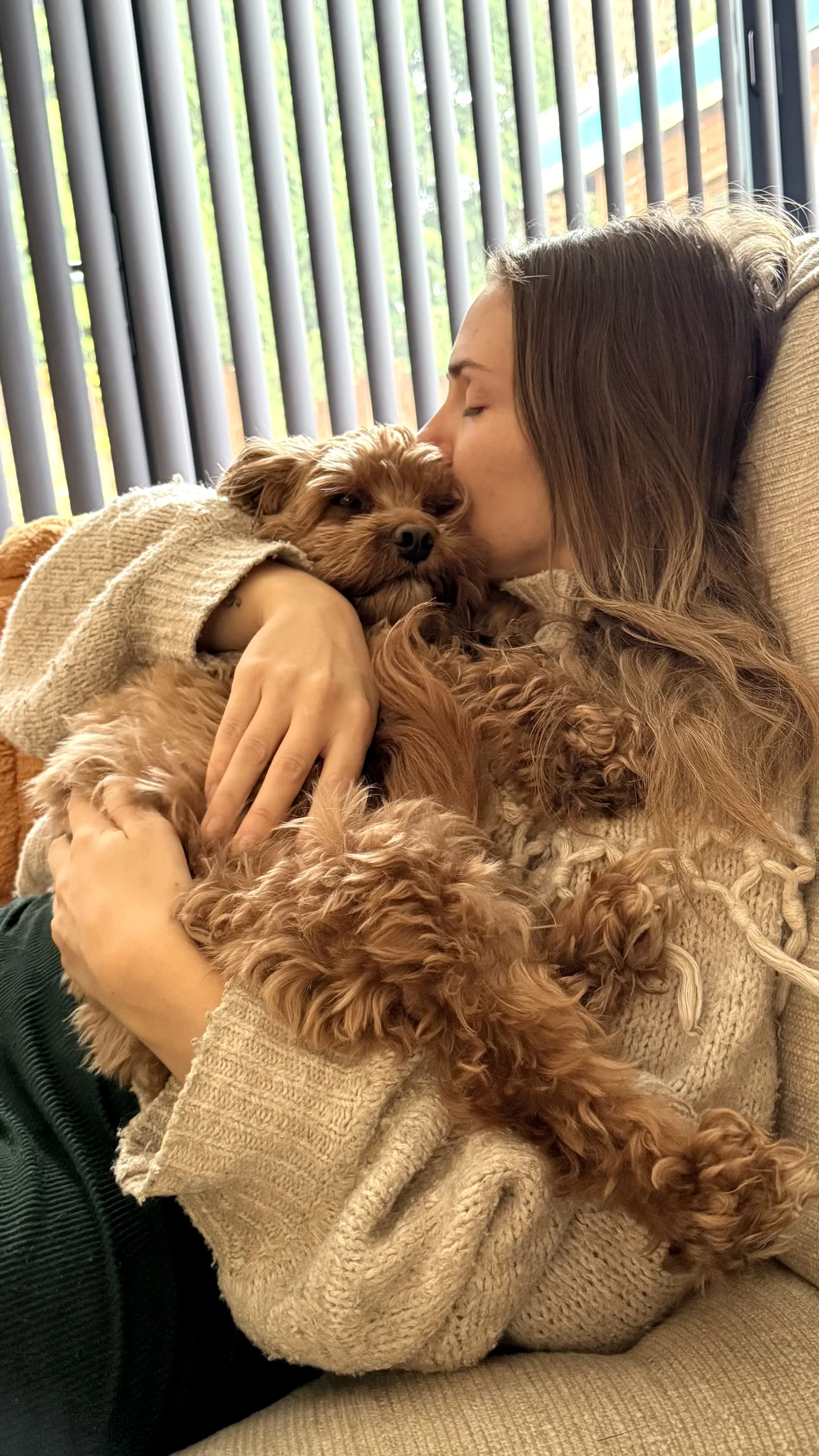 A woman with brown hair kisses a small, fluffy brown dog on the head while holding it in her arms on a couch. The woman is wearing a beige sweater, and the dog appears relaxed.