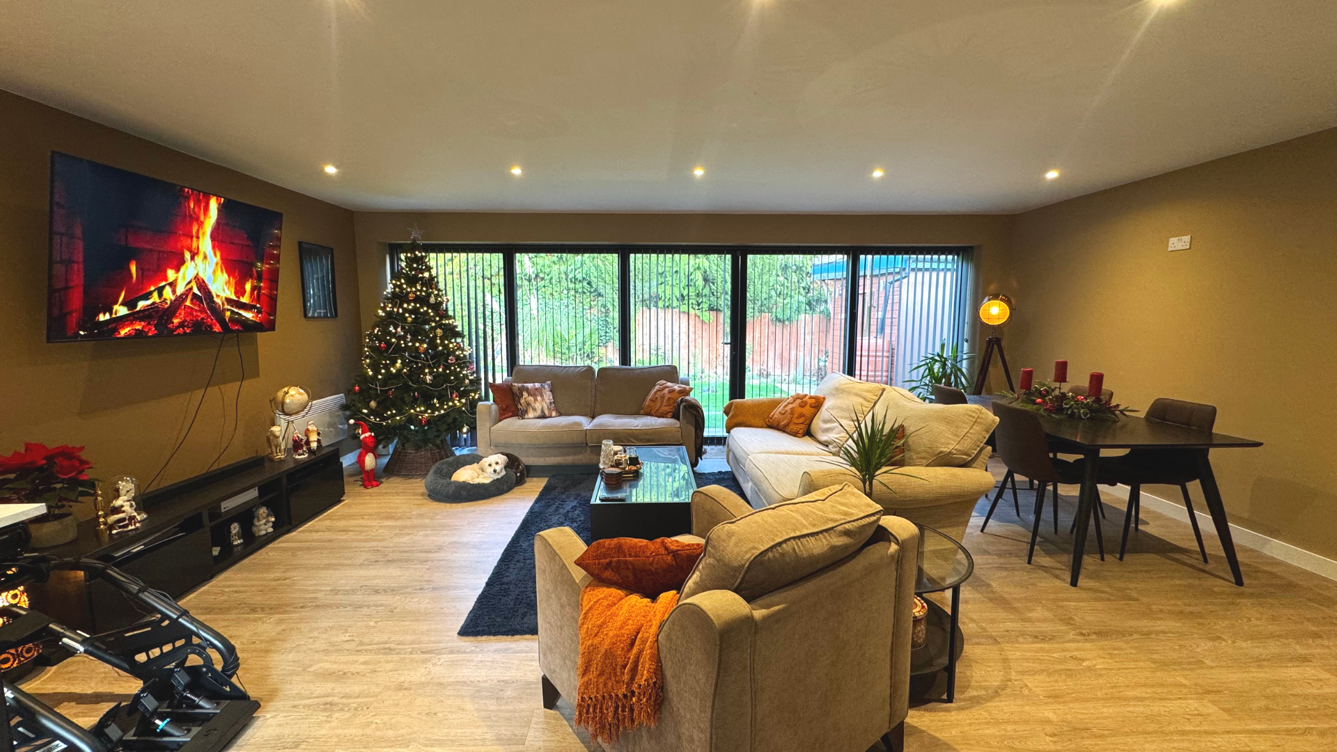 Living room with Christmas tree, holiday decorations, beige sofas, a black coffee table, a rug, a dining table with chairs, and sliding glass doors showing a backyard.