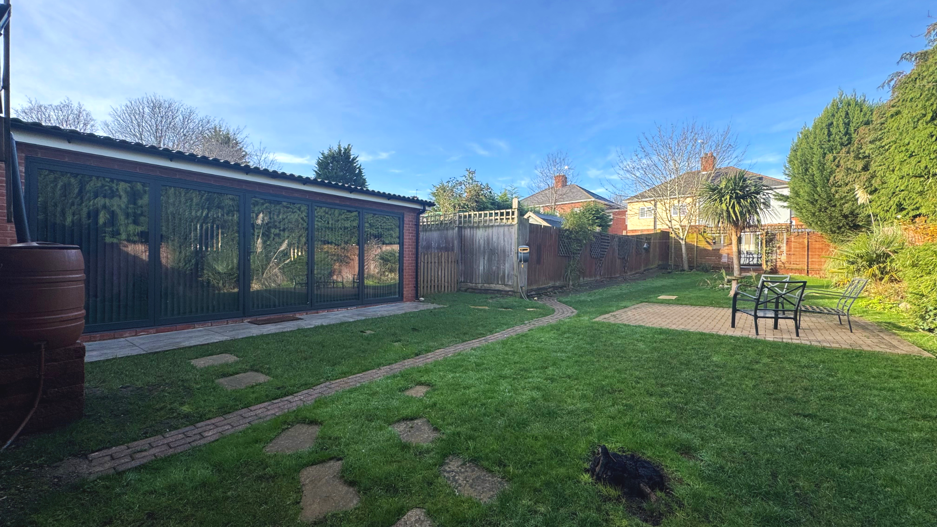 Backyard with grass, brick pathway, glass greenhouse, patio area with chairs, trees, and neighboring houses under a clear blue sky.