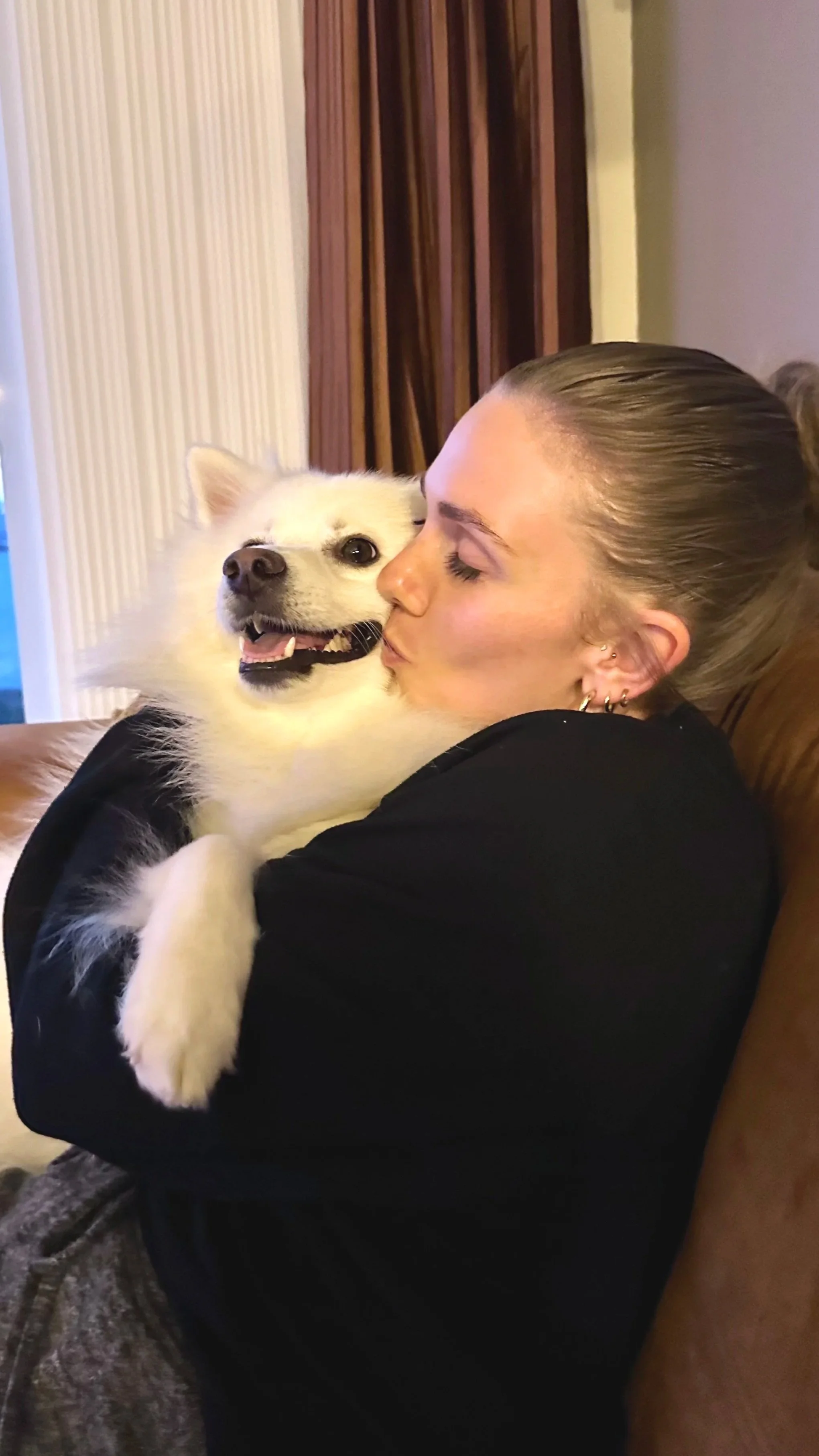 A woman with her eyes closed hugging and kissing a happy, fluffy white dog on the cheek.