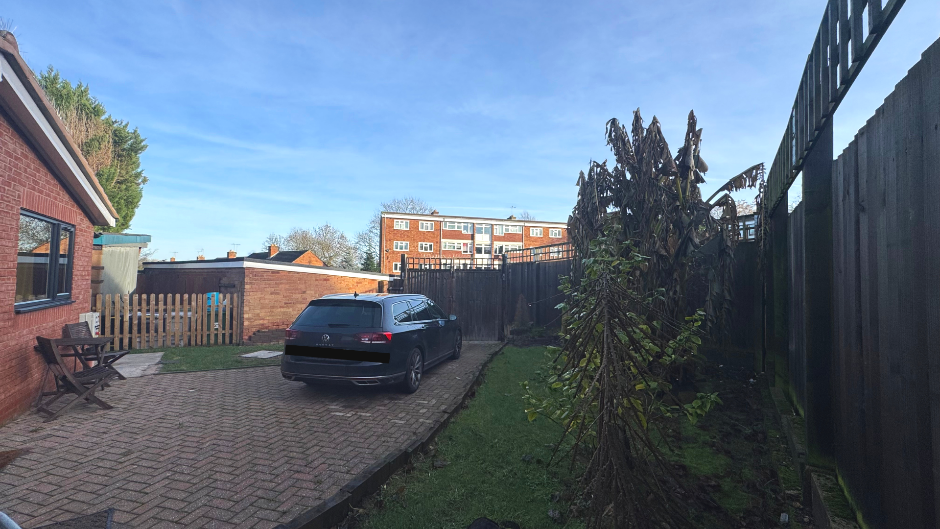 A backyard with a brick house on the left, wooden benches, a black station wagon, a small grassy area, a tall withered plant, and a tall wooden fence. In the background, there are multi-story brick buildings and a clear blue sky.