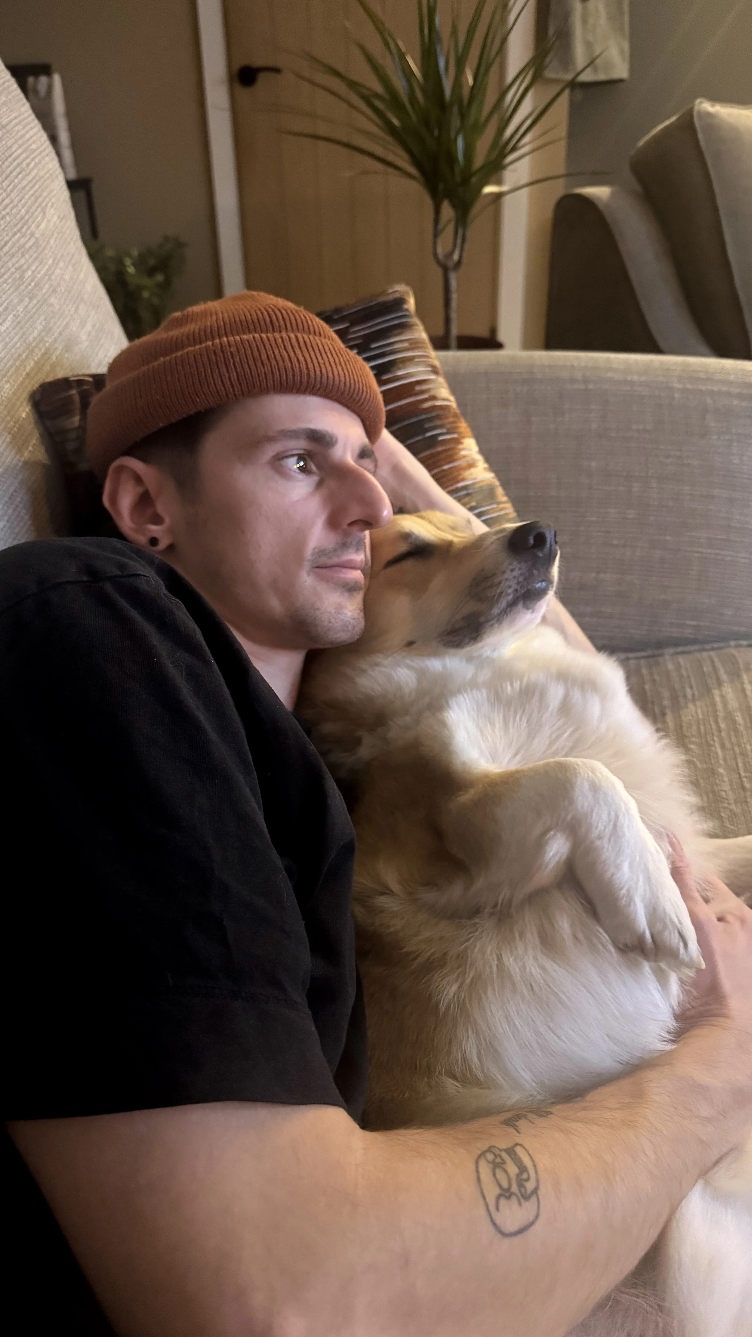 A man with a brown beanie and black shirt lounging on a couch with a corgi dog resting its head on his shoulder, both appearing relaxed and content.