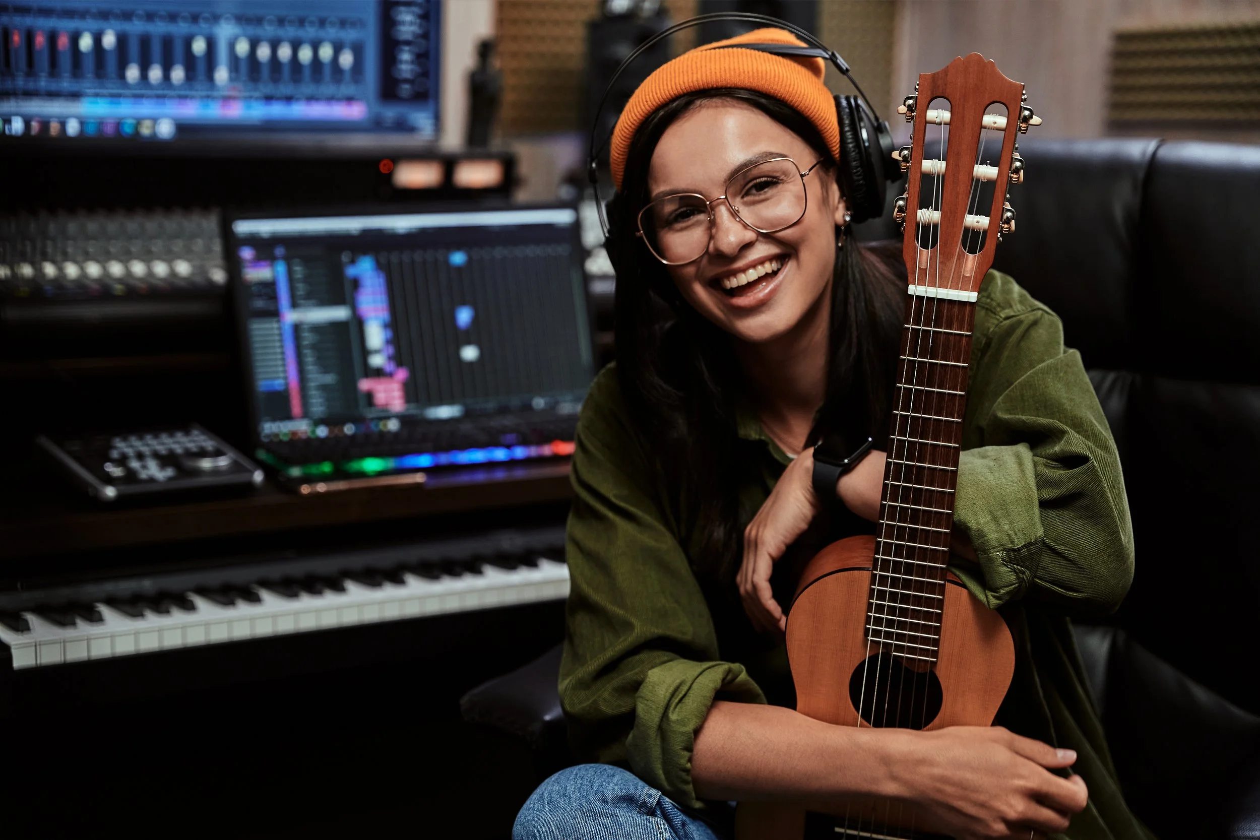 A smiling woman in headphones and glasses holding a guitar in a music studio with a keyboard, mixing console, and computer screens showing music editing software.