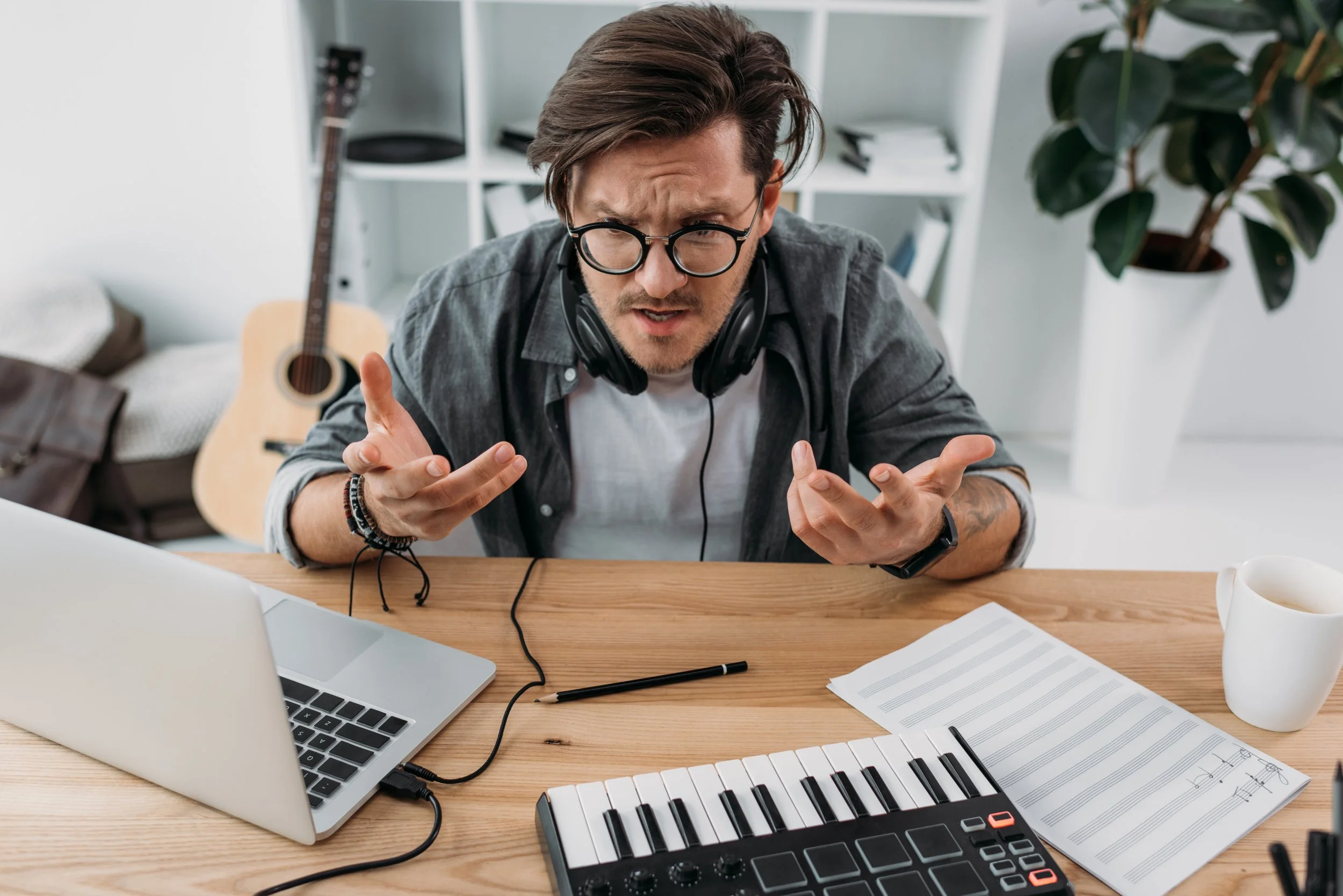 A young man wearing glasses, with headphones around his neck, sitting at a desk with a laptop, music keyboard, sheet music, and a coffee mug, gesturing with his hands while looking frustrated or confused in a home office setting.