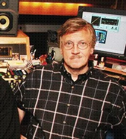 A man with glasses and a beard wearing a black and white checkered shirt, sitting in a music or recording studio with audio equipment and computer screens in the background.