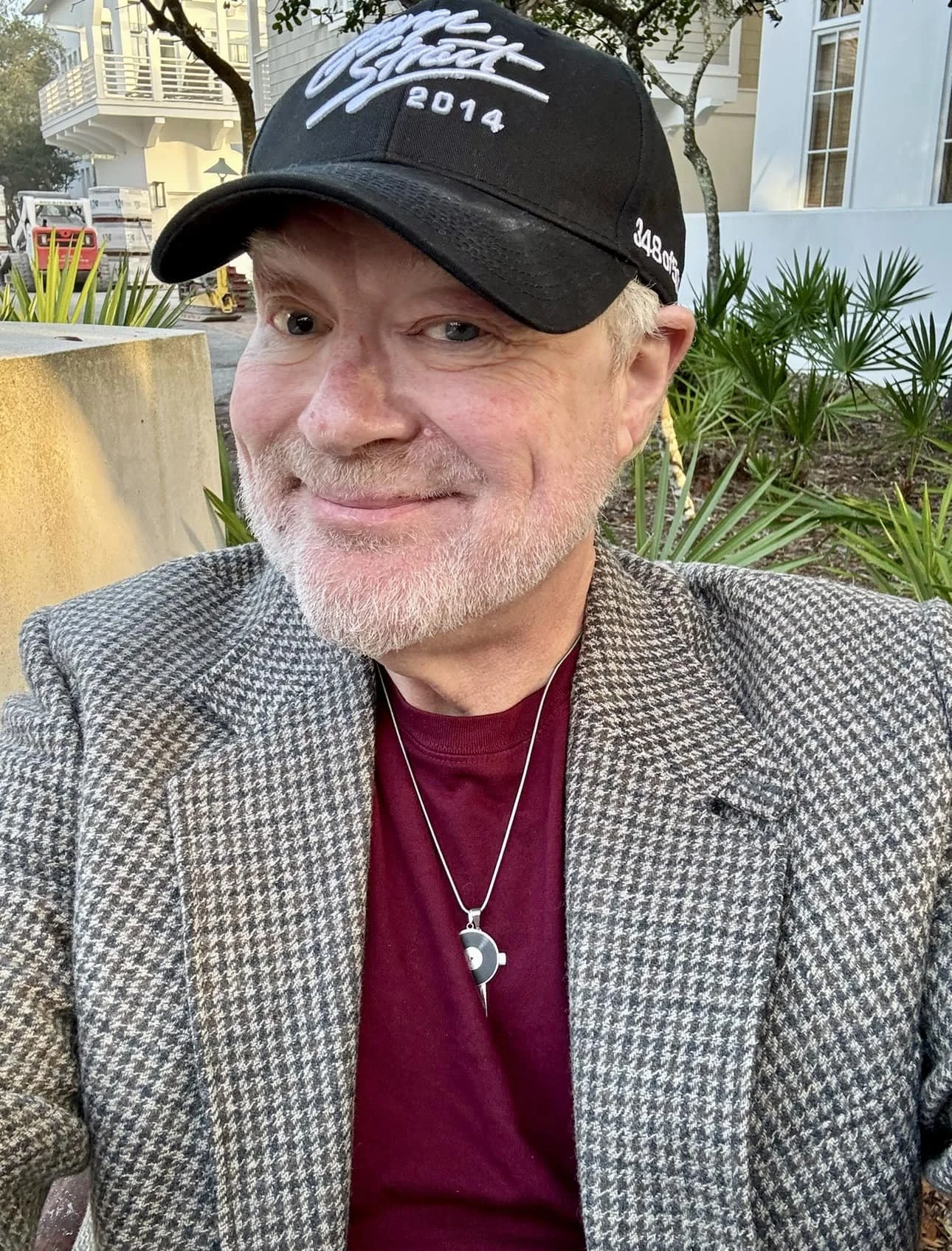 A middle-aged man with a gray beard and light hair taking a selfie outdoors. He is wearing a gray houndstooth blazer, a red shirt, a necklace with a yin-yang pendant, and a black baseball cap with white text and logo. Behind him are green plants, a tree, a building with white walls and balconies, and some outdoor equipment.