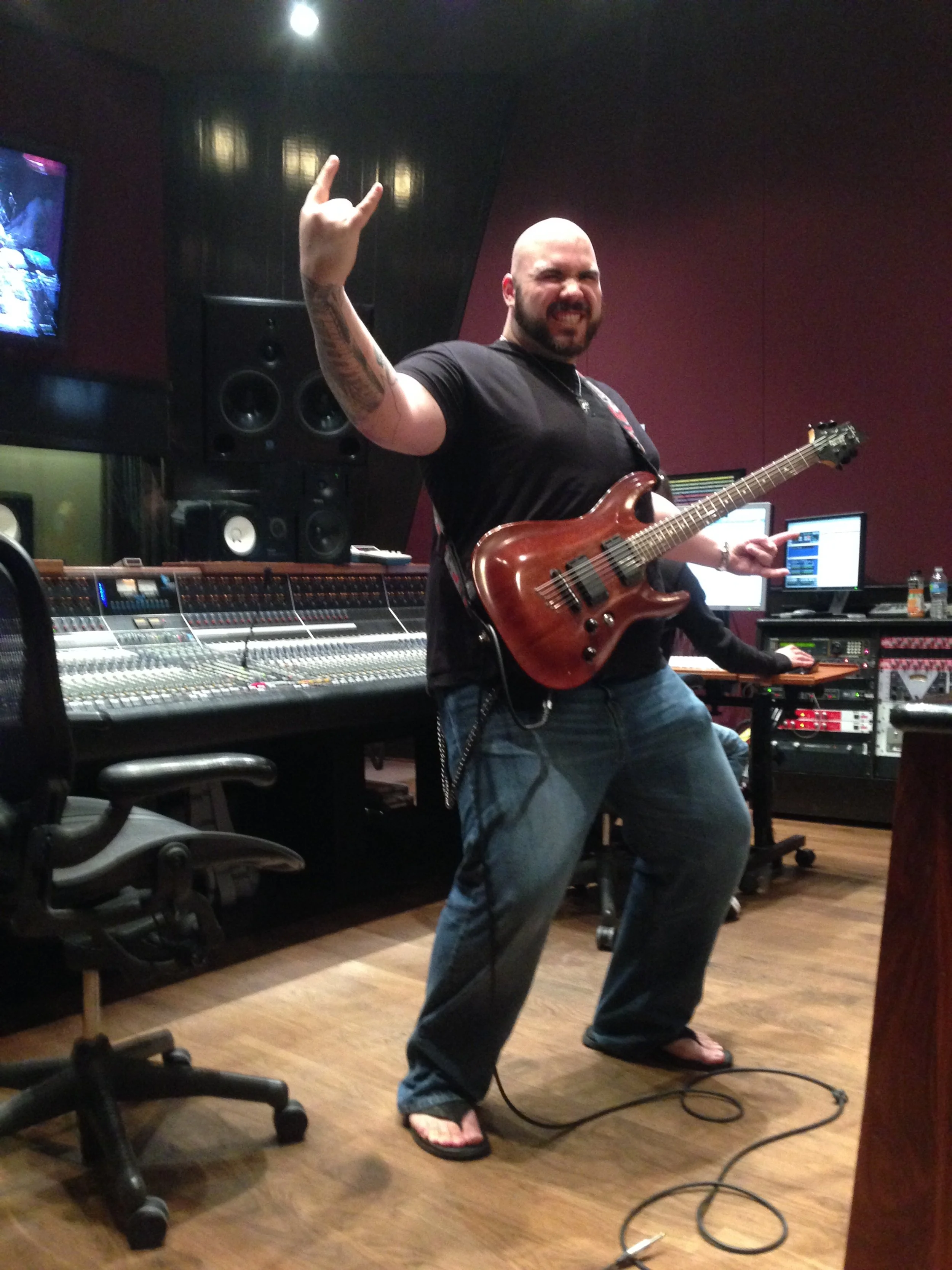 A man wearing a black shirt and jeans, holding an electric guitar and making a rock hand gesture, in a music studio with audio equipment and monitors.