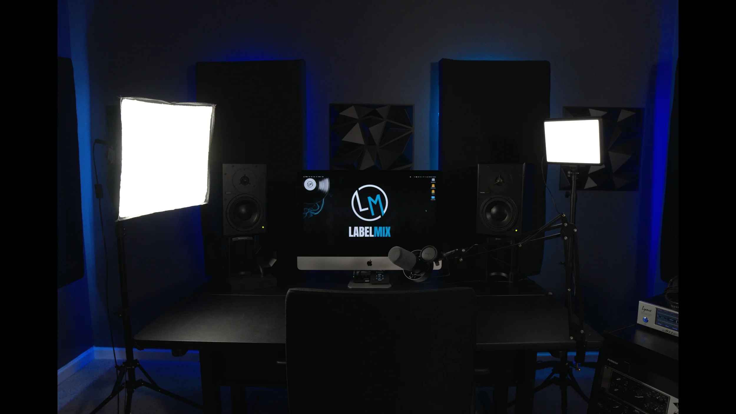 Recording or podcast studio setup with two bright LED lights, two studio monitors, a computer displaying 'LABELMIX', a microphone on a boom arm, and soundproofing panels on dark walls.