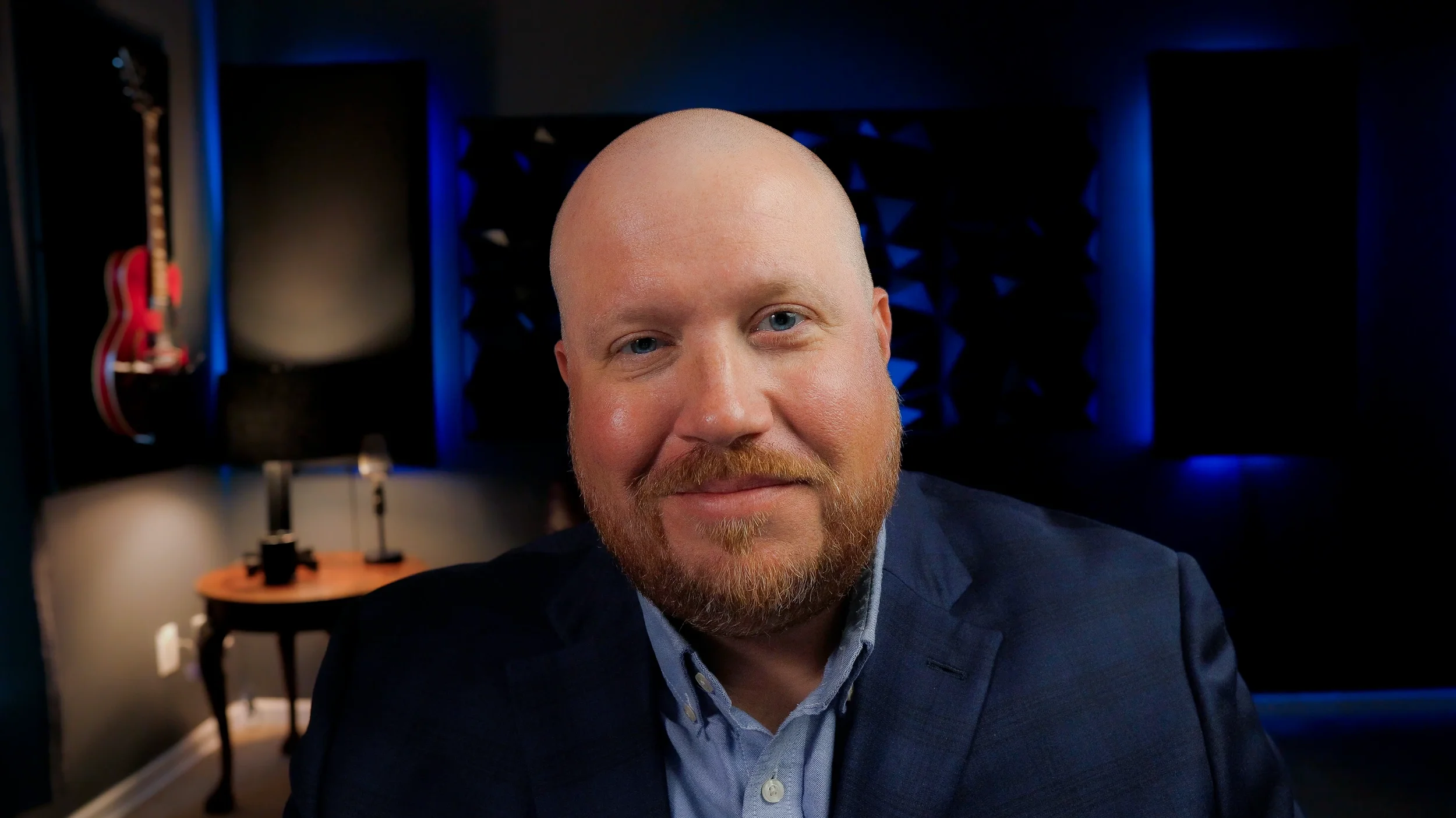 A man with a beard and mustache in a suit sitting in a recording studio with a guitar hanging on the wall behind him.